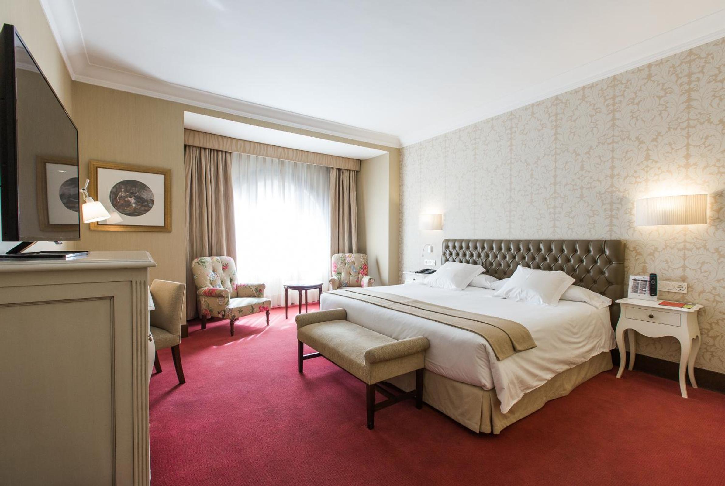 A hotel bedroom features a tufted headboard, red carpet, and floral armchairs beside a draped window.