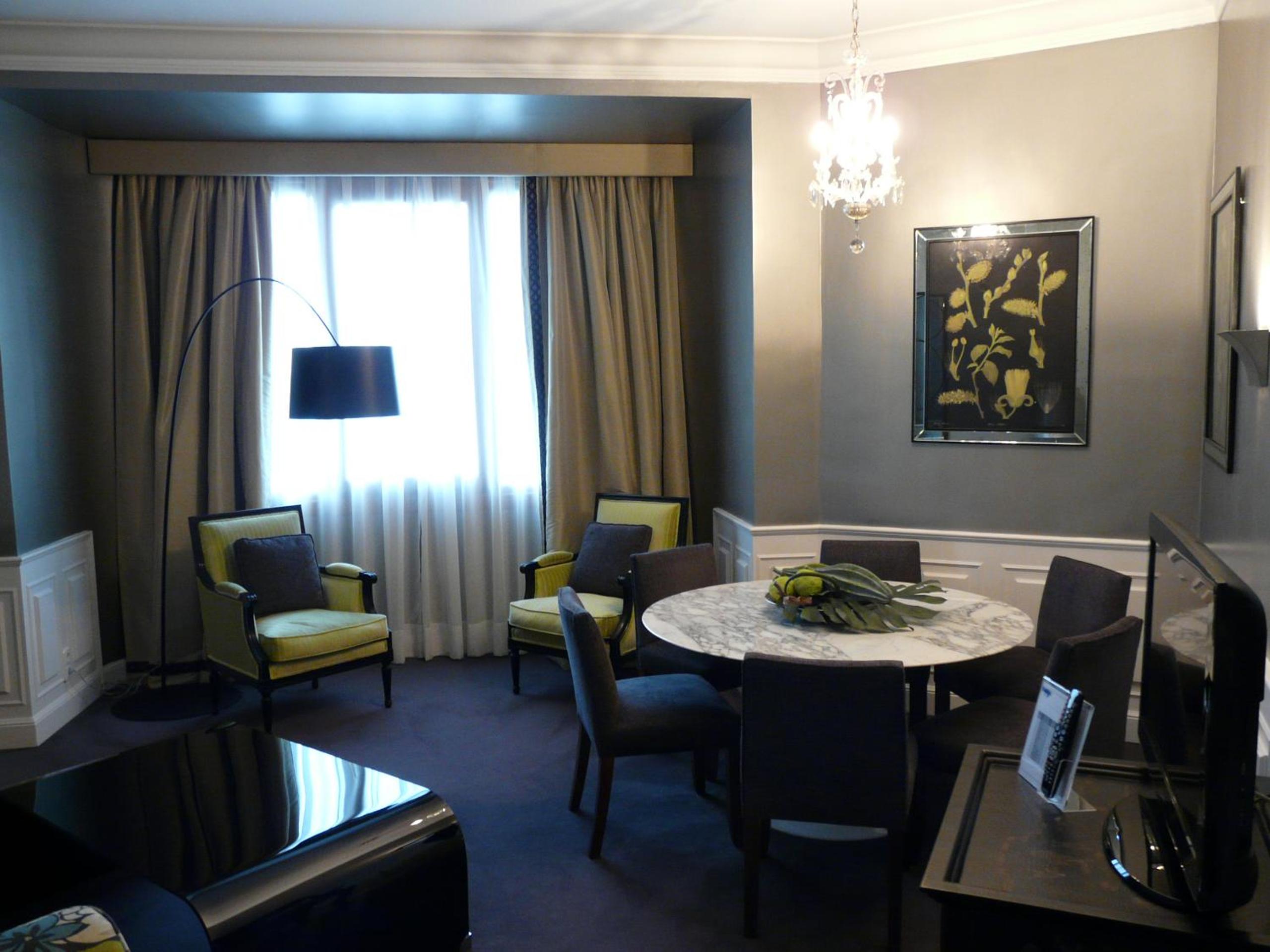 A grey sitting room contains a round marble table, dark chairs, armchairs, and a crystal chandelier.
