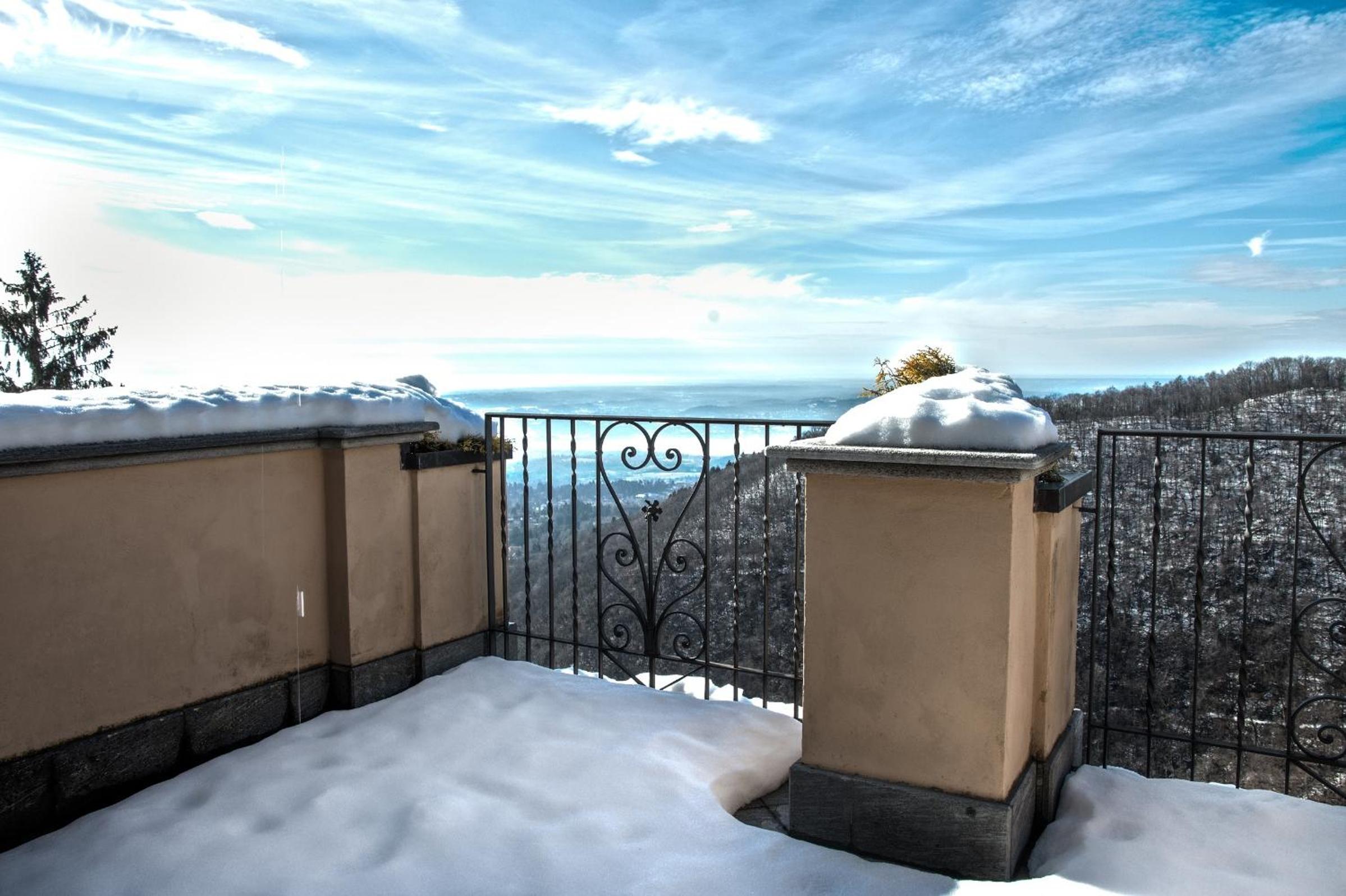 A snow-covered balcony with ornate iron railings overlooks a distant valley and winter hills under a bright blue sky.