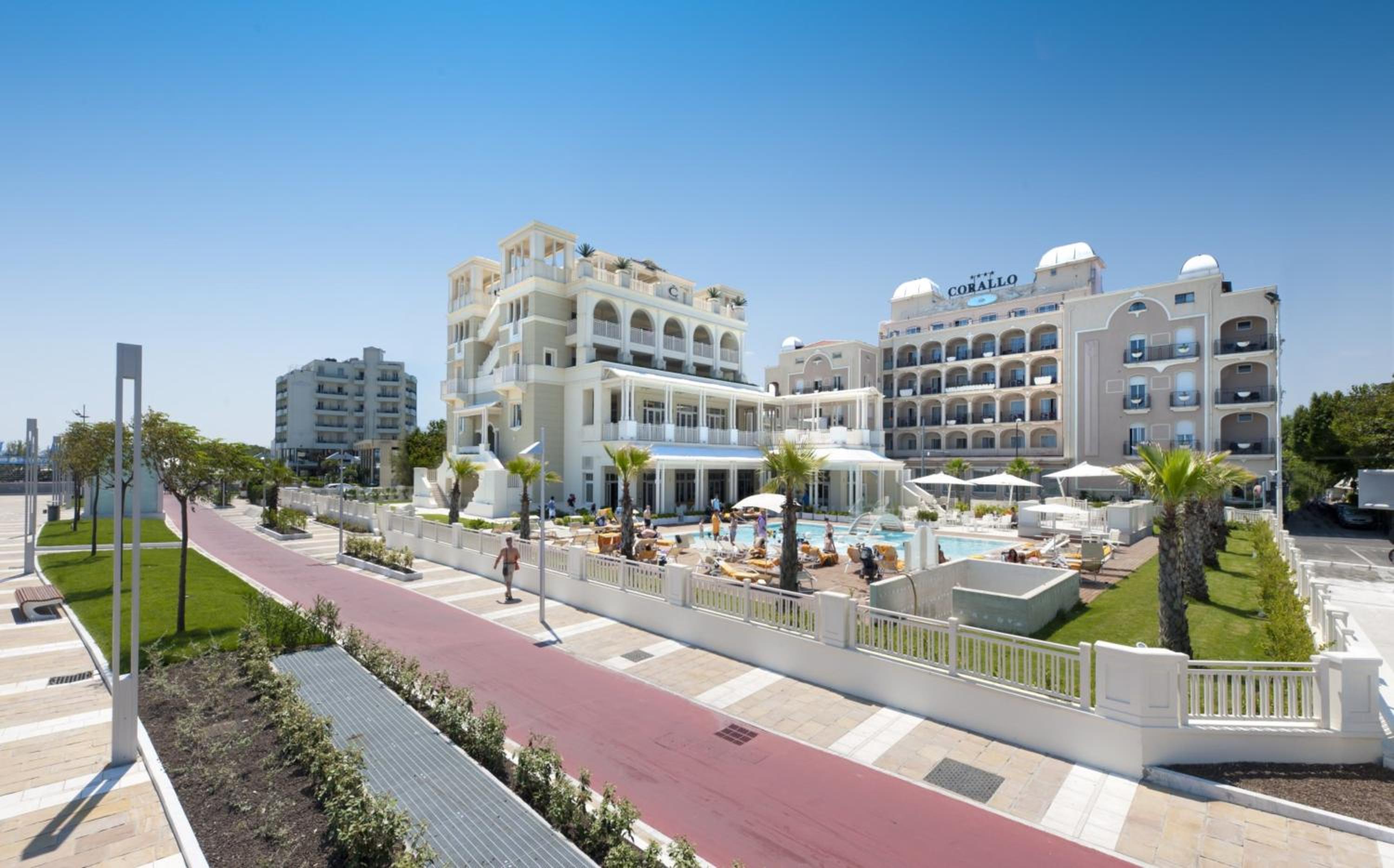 Exterior view of Hotel Corallo with an outdoor pool, sun loungers, palm trees, and a paved walkway under a clear sky.