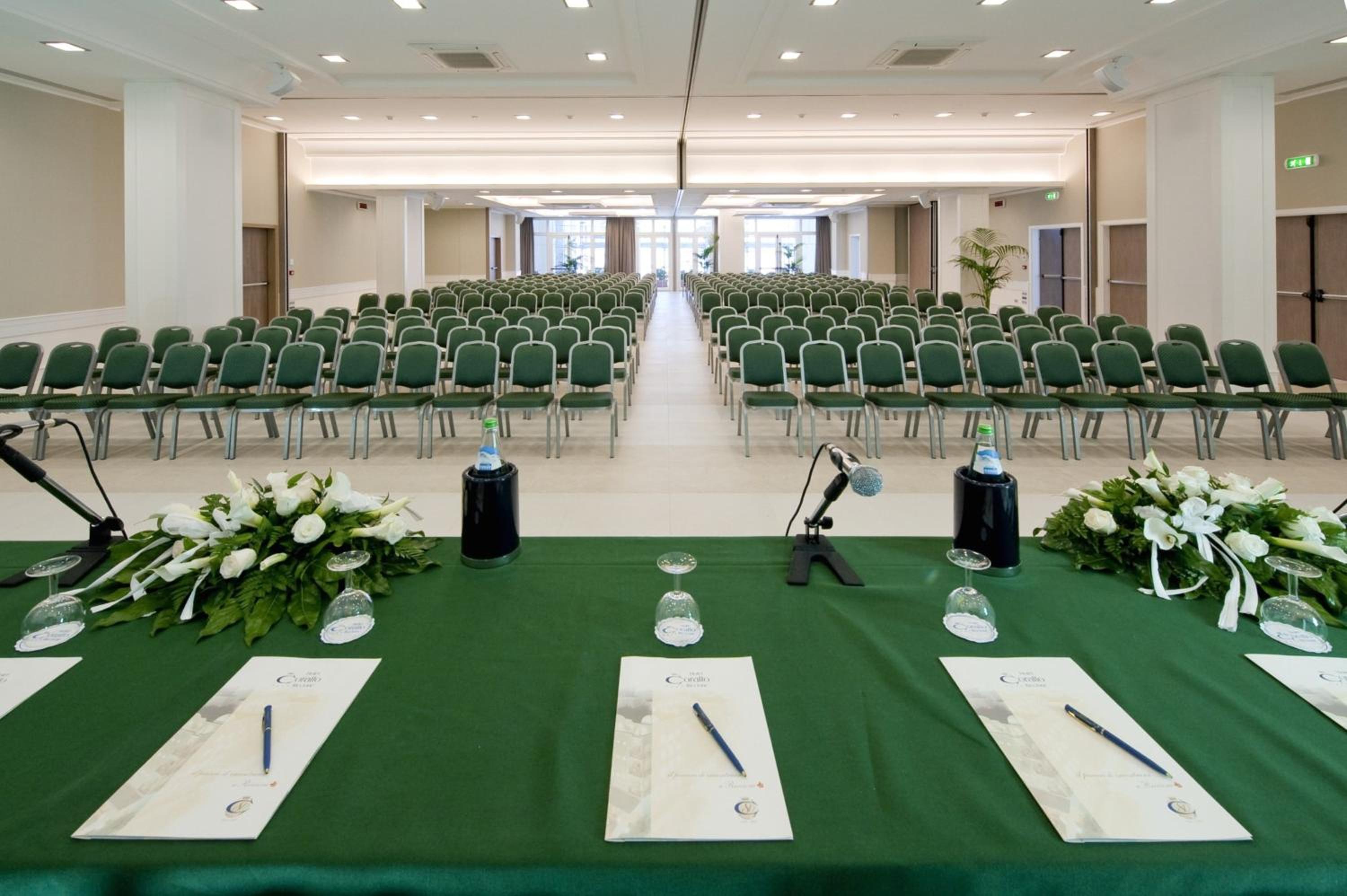 Large conference room with rows of green chairs facing a presentation table decorated with white flowers.