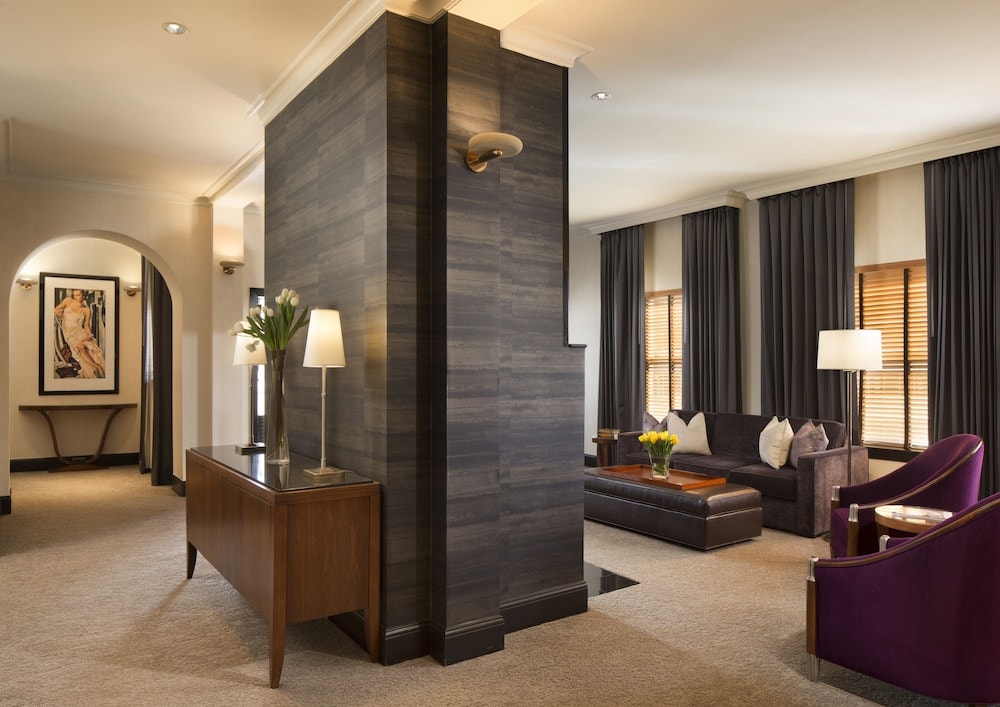 A hotel suite living area with a dark textured central wall, plush sofa, purple armchairs, and neutral carpet.