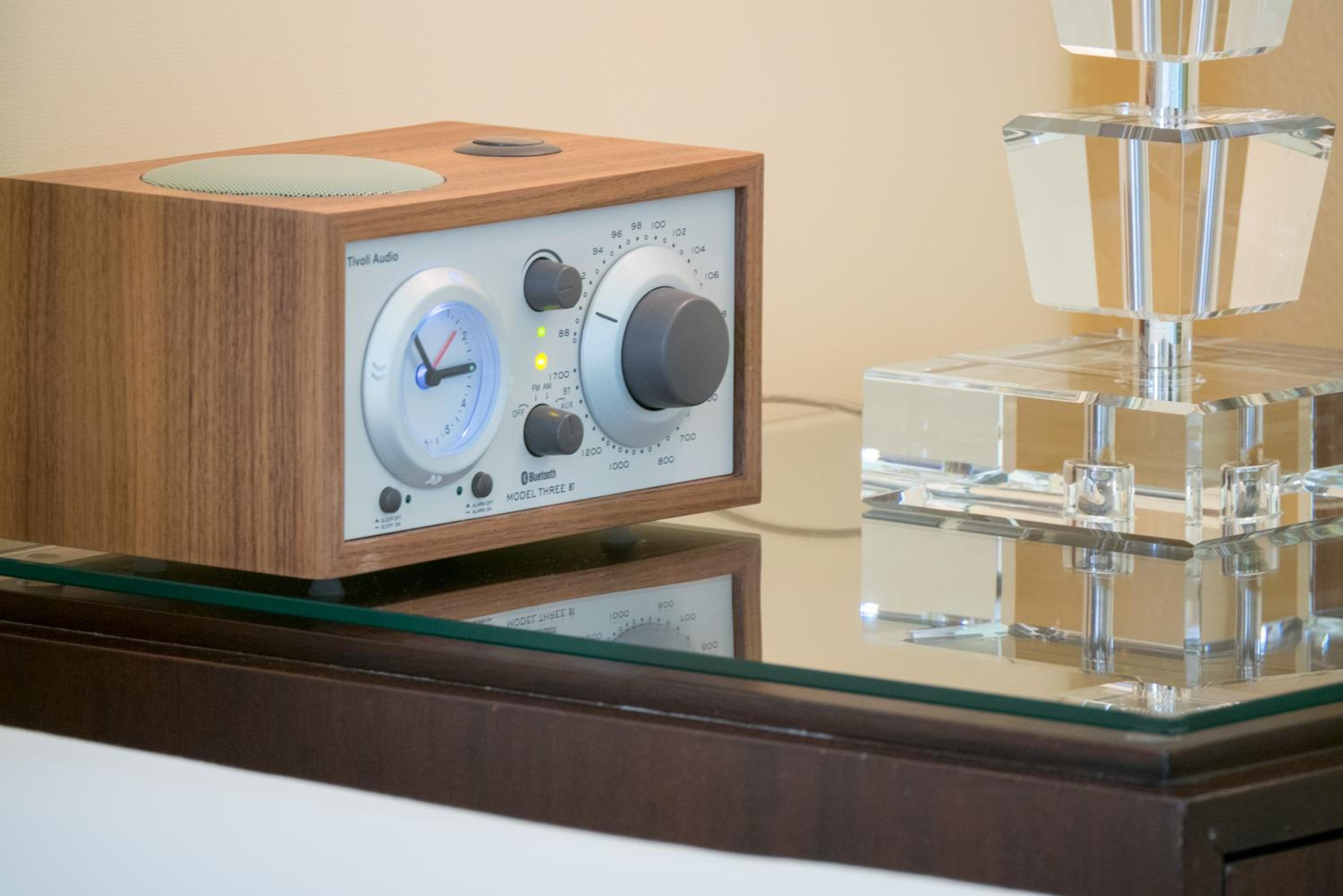A wooden radio alarm clock and crystal lamp base on a glass-topped nightstand.