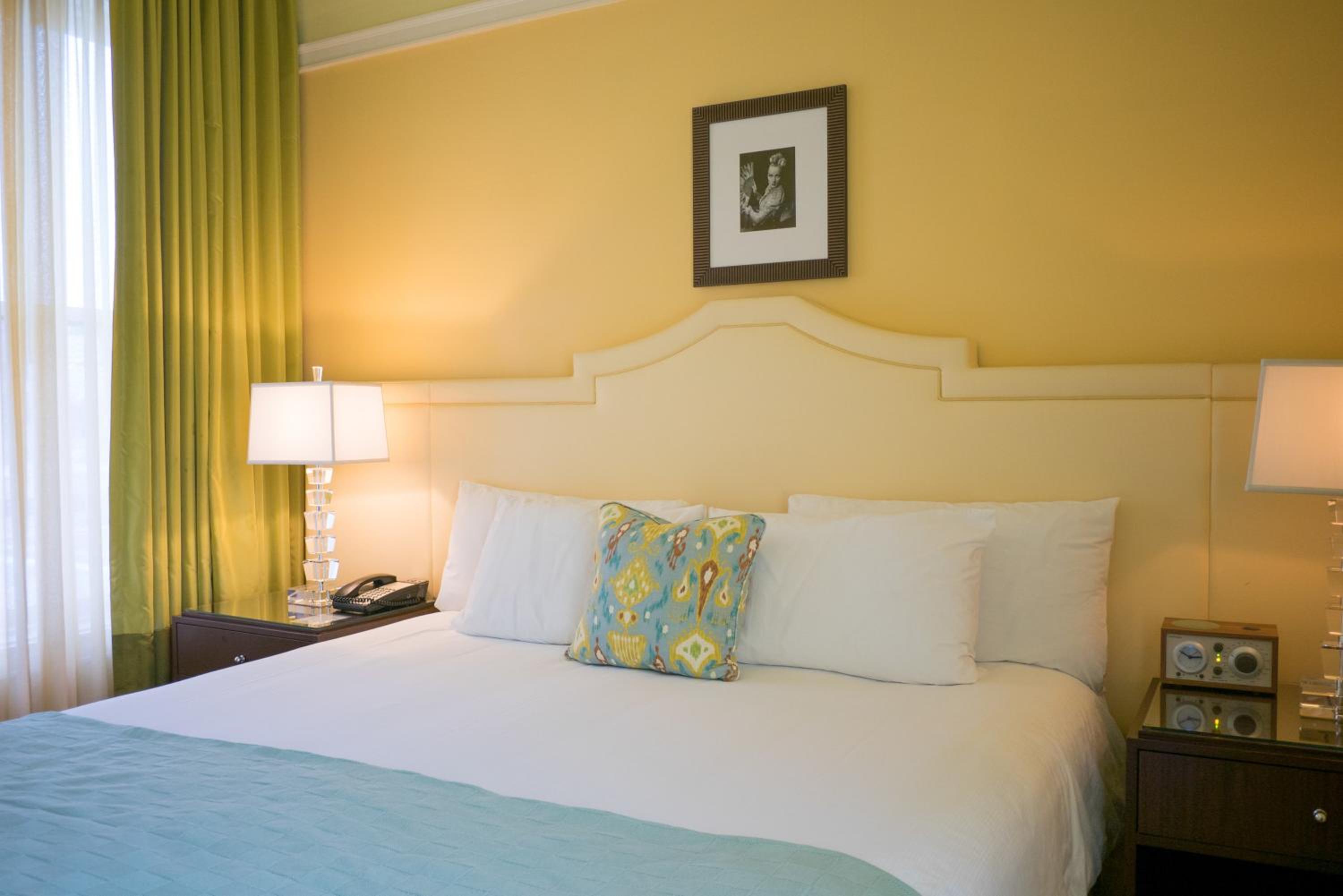 A hotel bedroom with a white upholstered headboard, yellow walls, green curtains, and framed wall art.