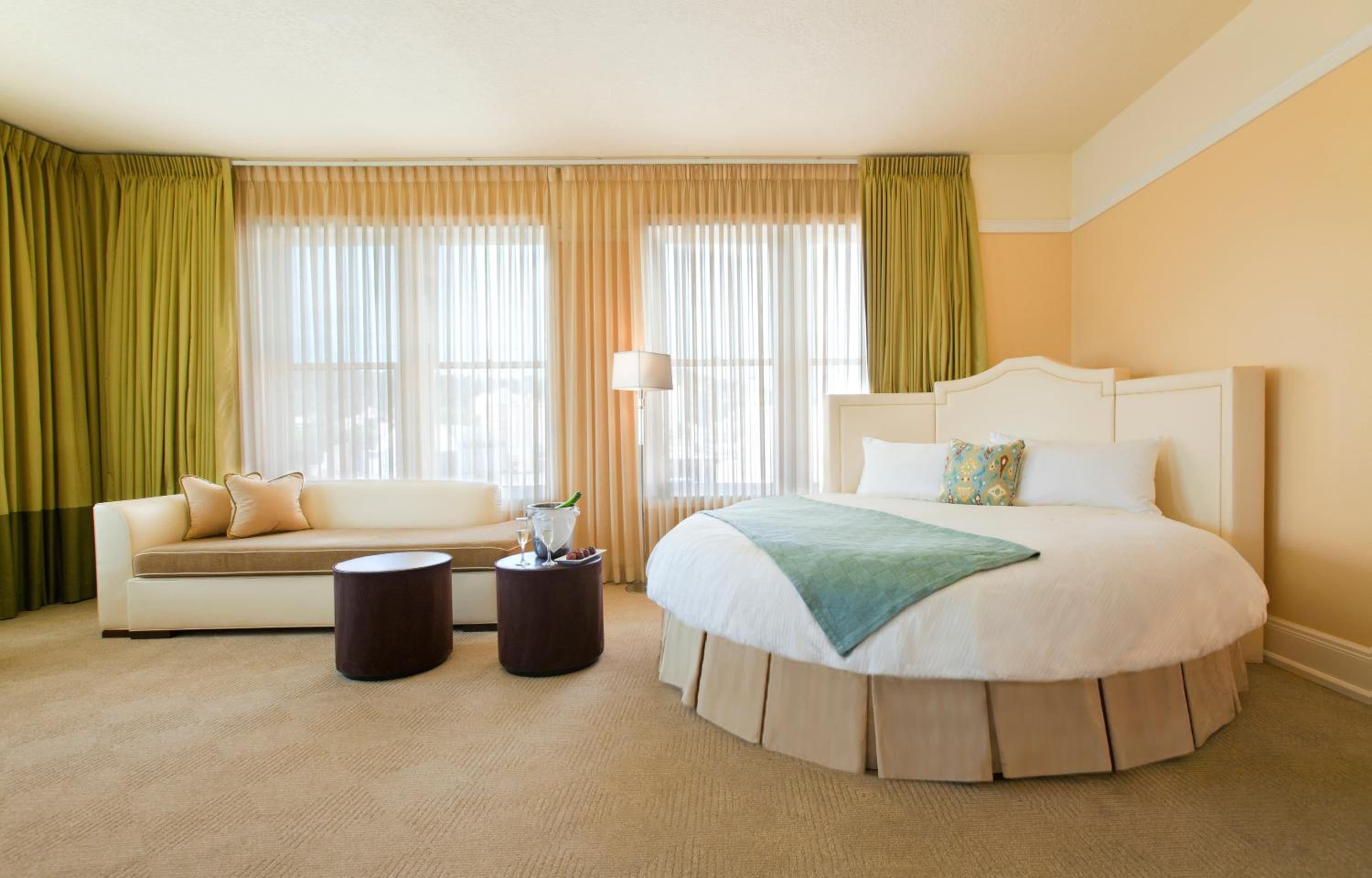 A hotel suite featuring a round bed, a daybed sofa, and large windows with sheer drapes.