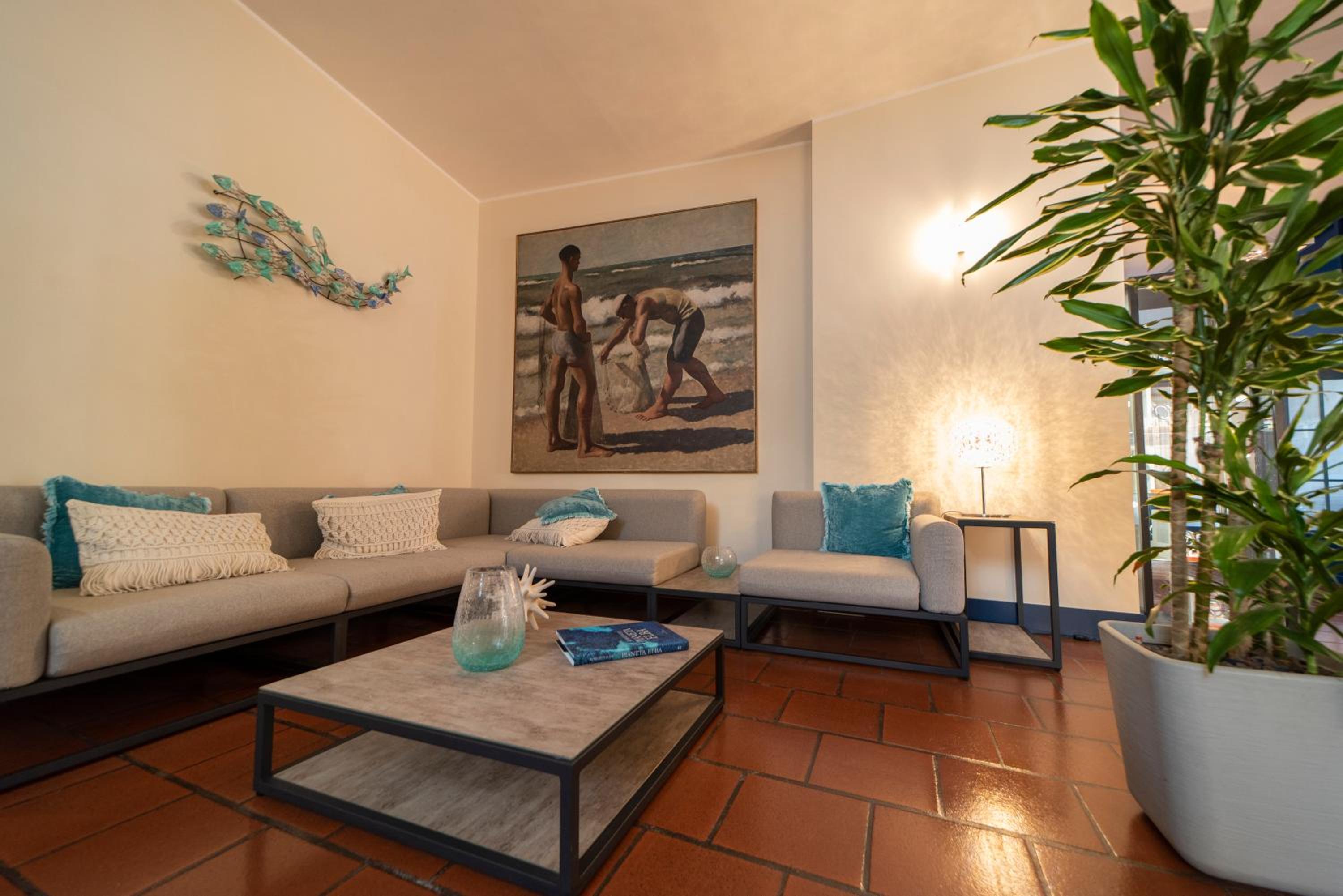 A hotel lounge with terracotta floor tiles, gray sectional sofas, a potted plant, and a beach painting.