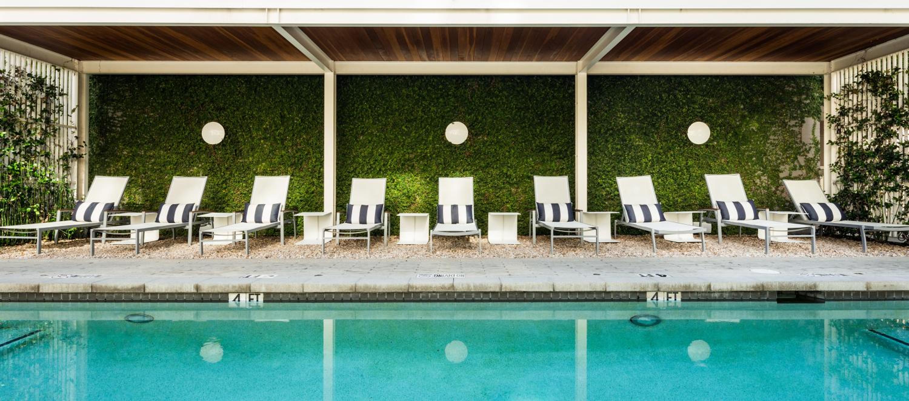 Turquoise swimming pool bordered by lounge chairs under a pergola against an ivy wall.