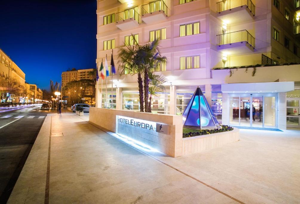 Illuminated exterior facade and entrance of Hotel Europa at night, with palm trees and a glowing glass pyramid.