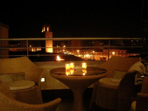 Wicker armchairs around a candlelit table on a rooftop terrace overlooking an illuminated town tower at night.