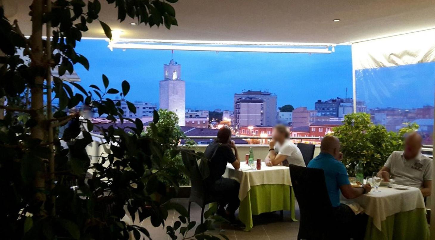 Guests dining at tables on a covered terrace overlooking city rooftops and a civic tower at dusk.