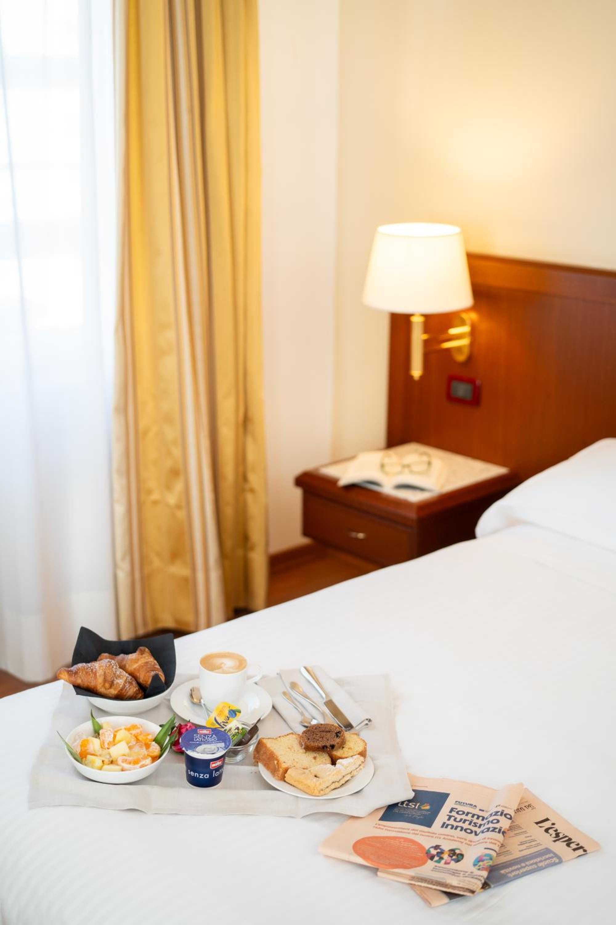 A breakfast tray with pastries, coffee, and fruit set on white bedding beside a wooden nightstand.