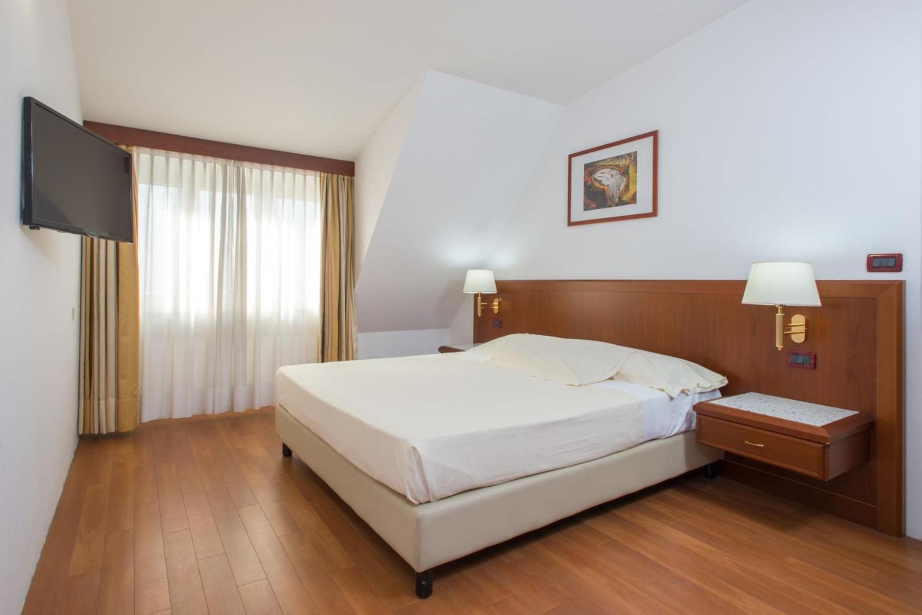 A bedroom with wood floors, a wooden headboard, bedside lamps, and sheer curtains covering a window.