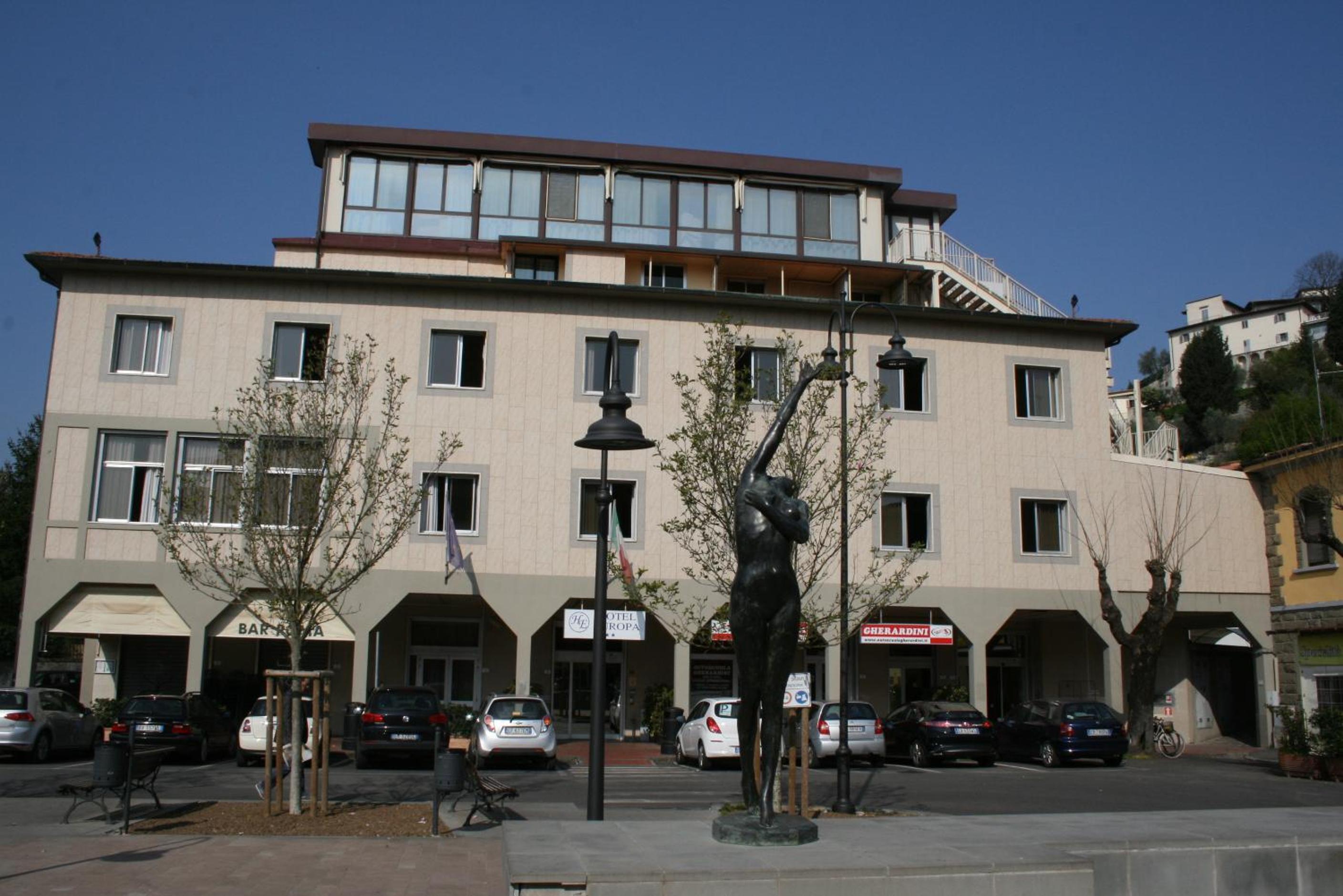 A dark bronze statue stands on a paved square before the multi-story facade and parking area of Hotel Europa.