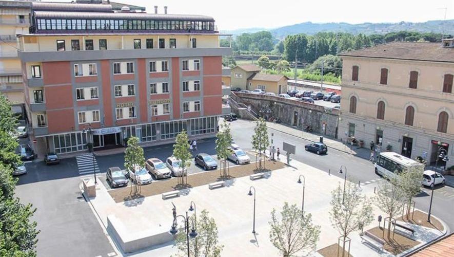 An elevated view looks over a paved town square with young trees, parked cars, and multi-story streetfront buildings.