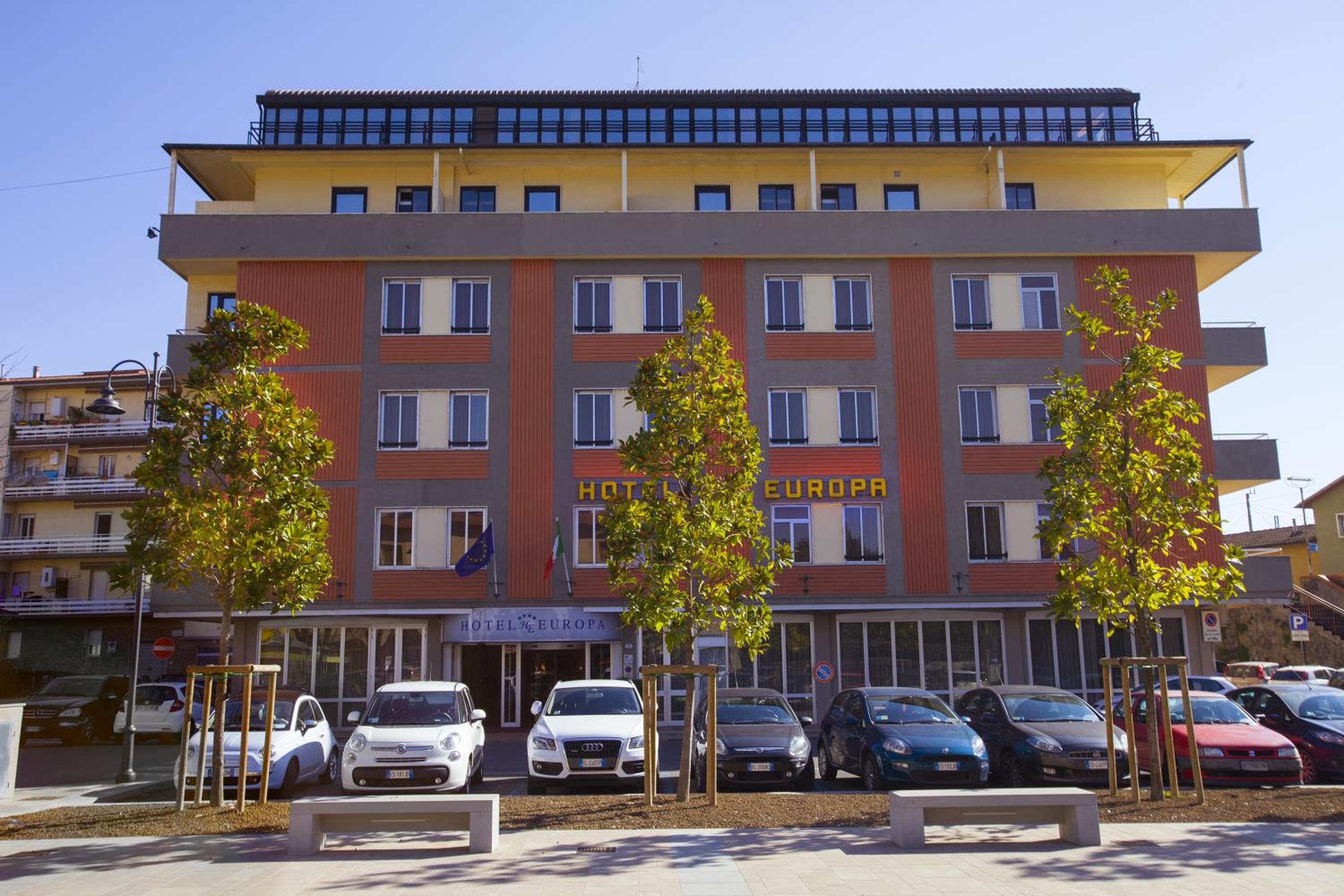 The exterior facade features orange panels, a yellow upper floor, and rows of parked cars fronted by slender trees.