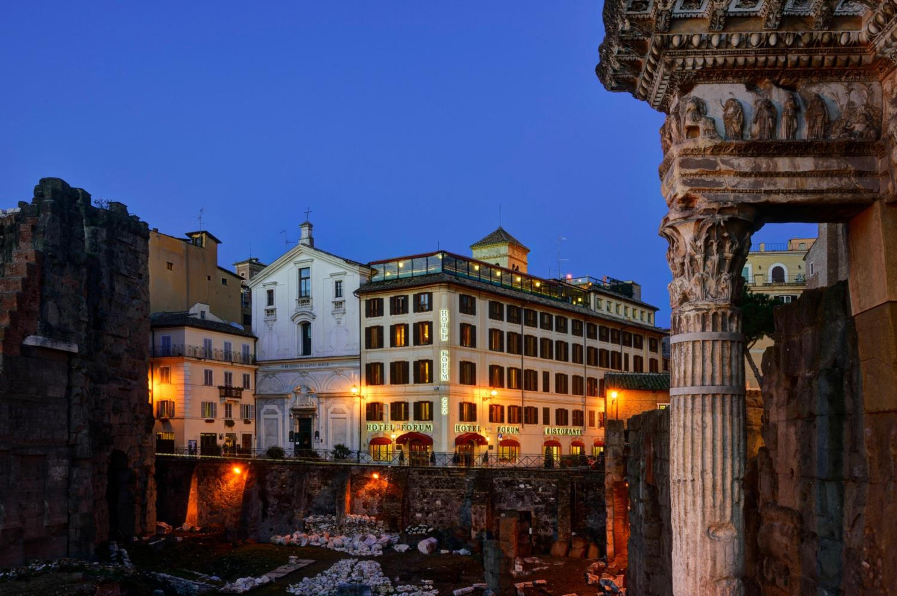 Illuminated facade of Hotel Forum at dusk, viewed past ancient Roman stone ruins and a carved column.