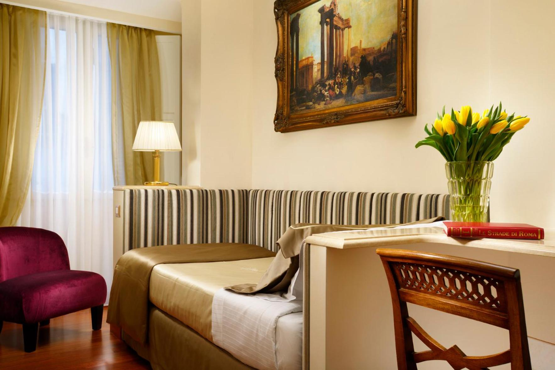 A daybed with striped upholstery sits beneath a gilded painting, beside a vase of yellow tulips.