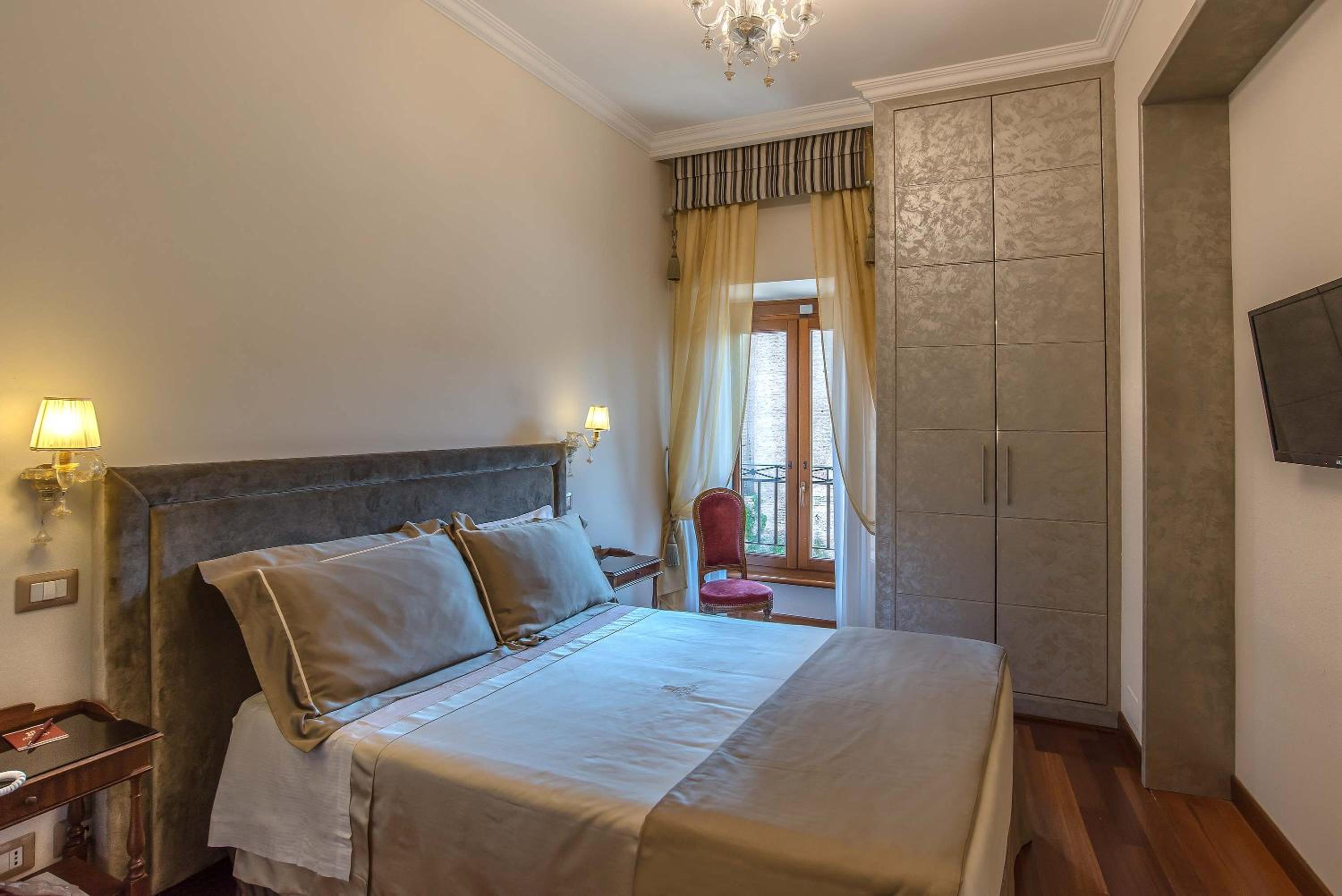 A bedroom with an upholstered bed, tall wardrobe, wood floor, and sheer curtains over a window.