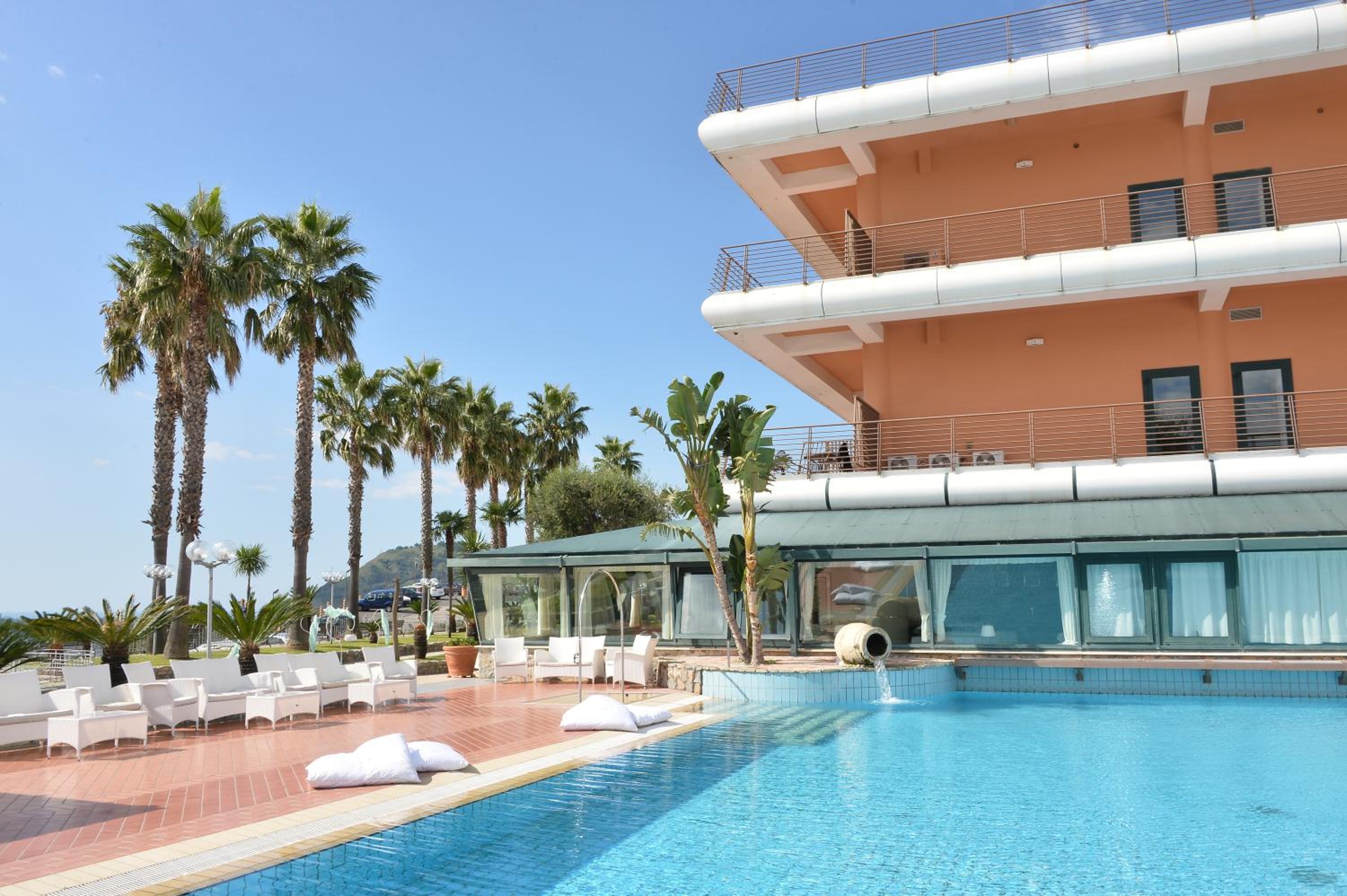 Swimming pool alongside a terracotta-colored building and palm trees under clear daylight.