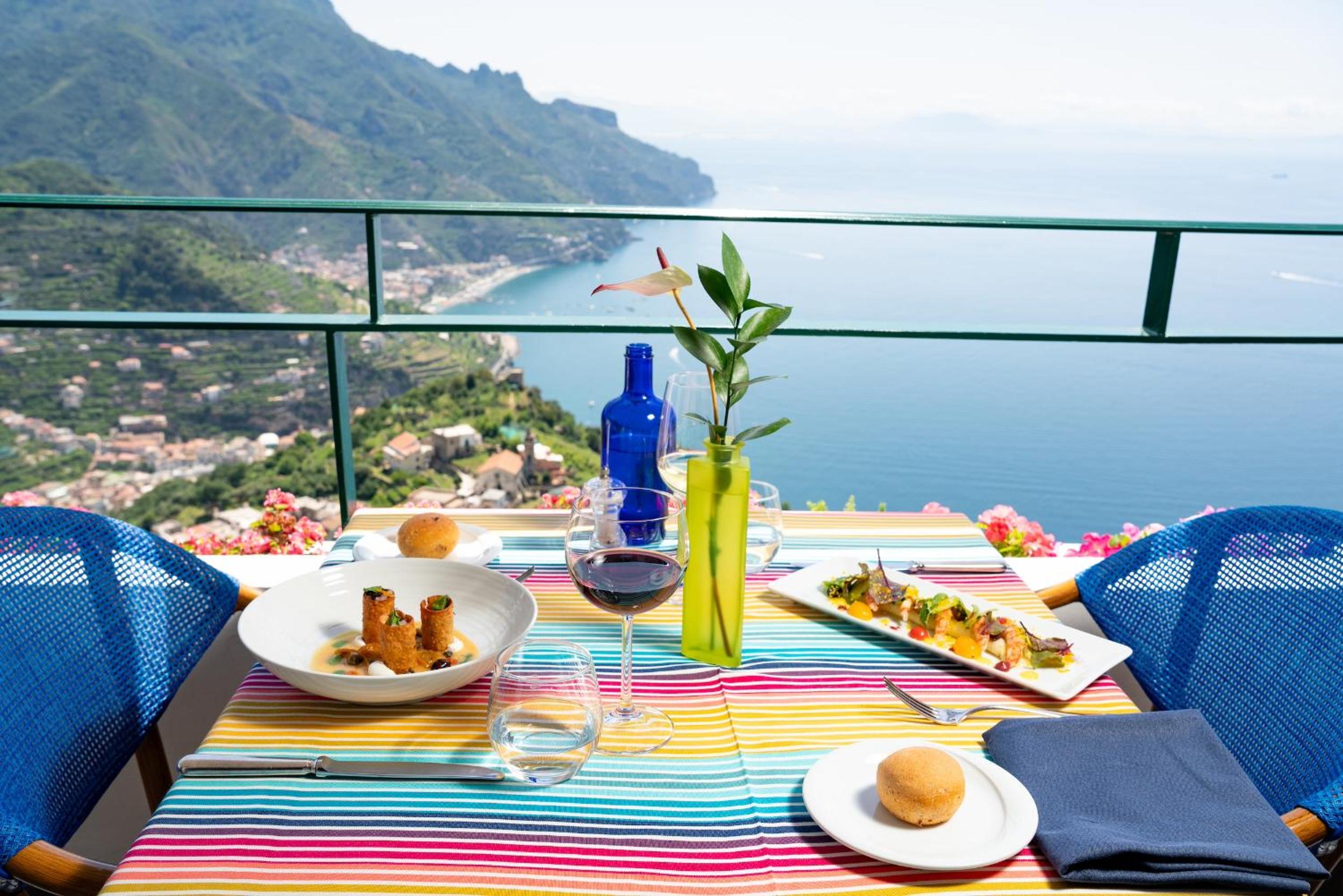 A table set with plated food and wine on a balcony overlooking the coastline and sea.