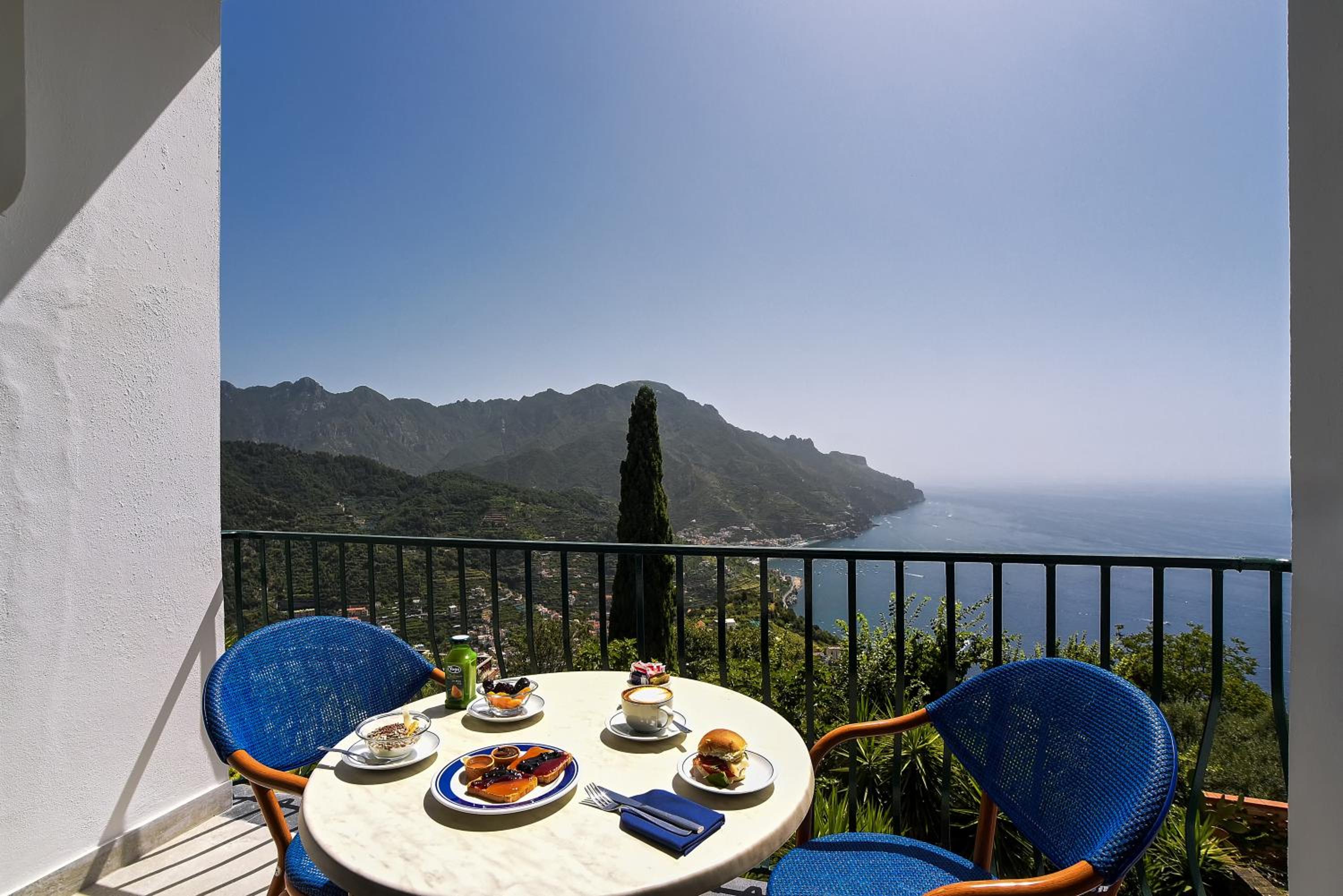 Breakfast spread on a balcony table with woven chairs facing coastal mountains and the sea.