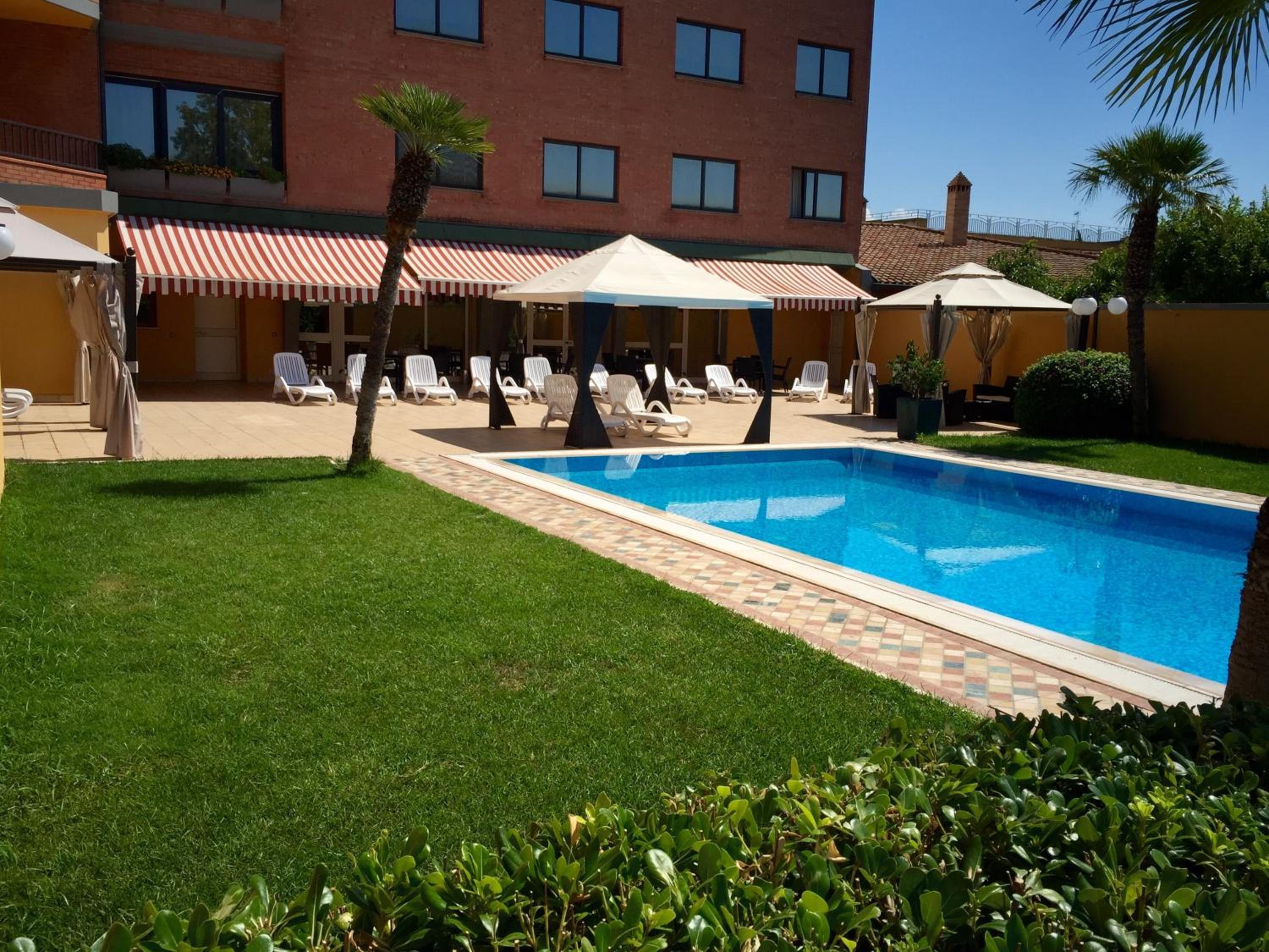 An outdoor swimming pool bordered by green lawn, lounge chairs, and shaded gazebos beside the building.