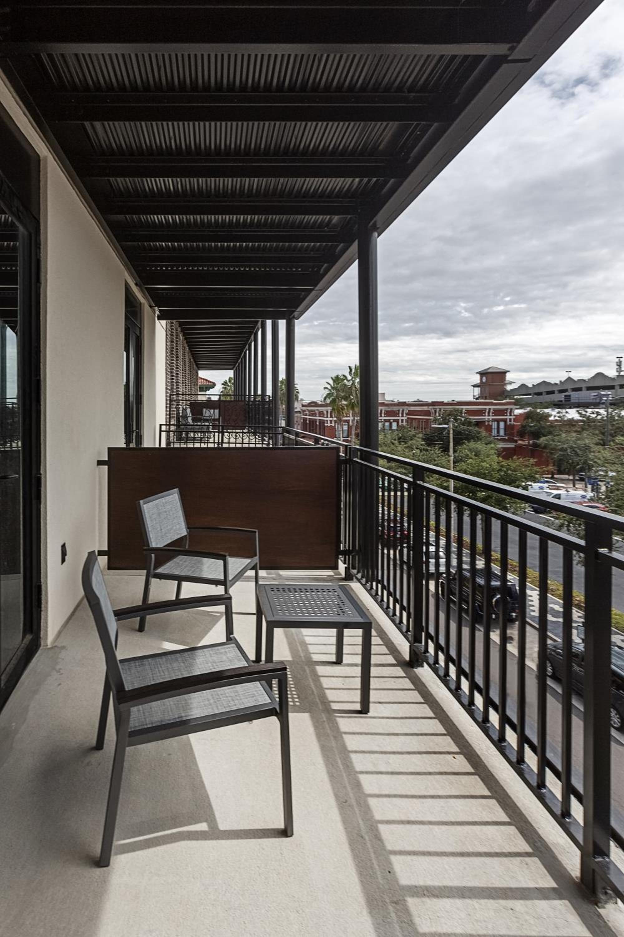 A covered balcony with metal chairs and a small table overlooking street traffic and brick buildings.