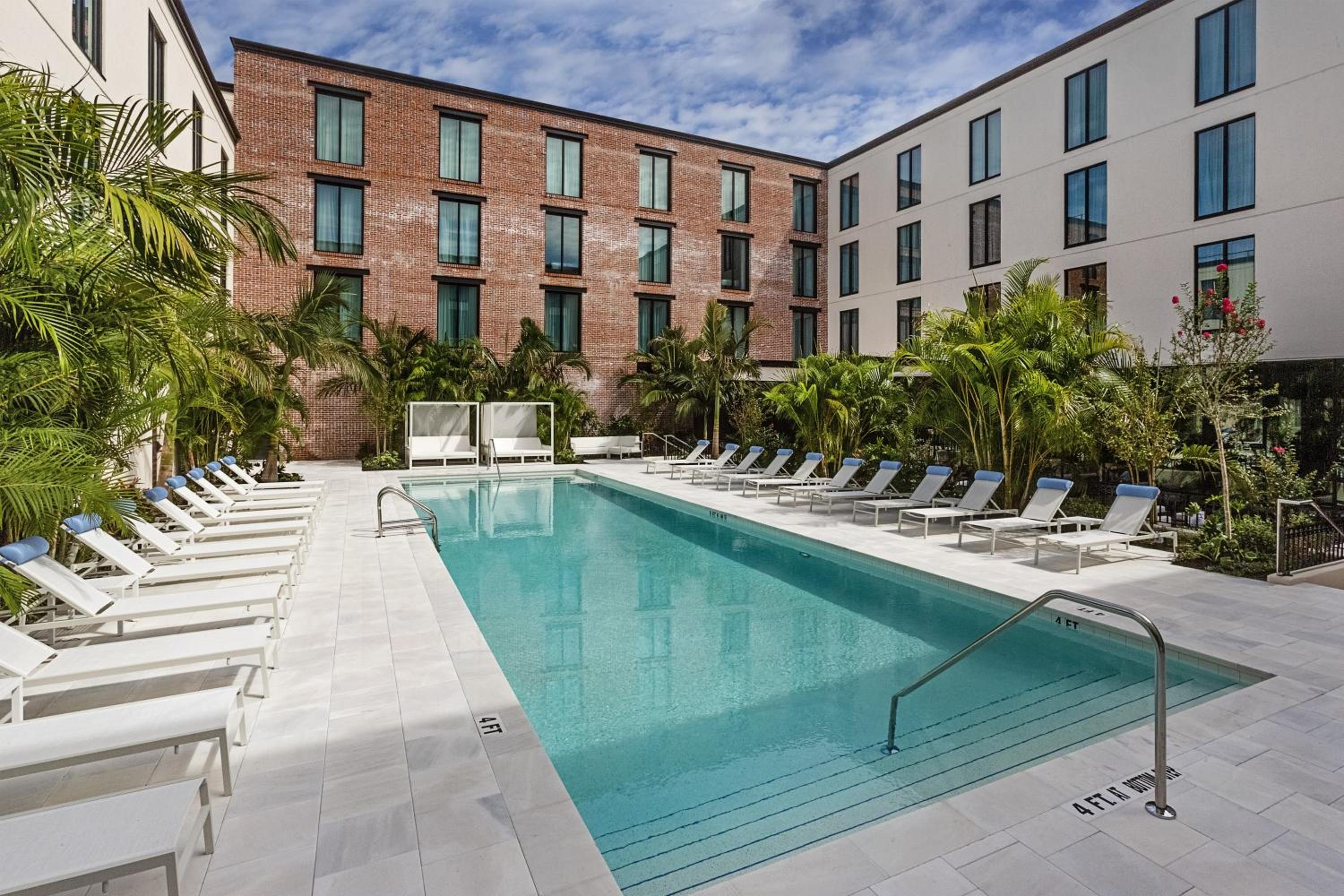 A courtyard swimming pool surrounded by white lounge chairs, palm trees, and multistory brick walls.