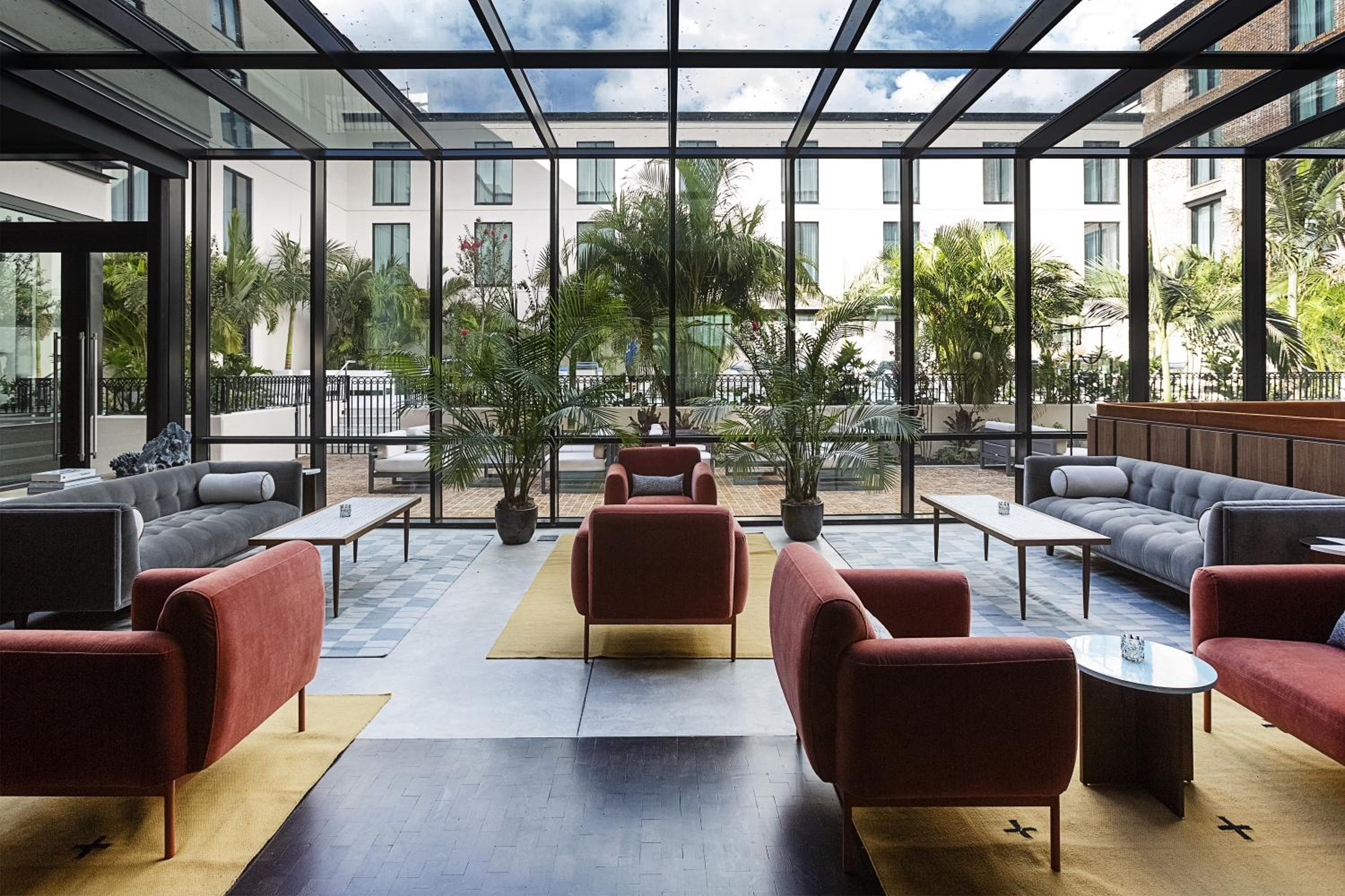 A glass-enclosed lounge with skylights, armchairs, and sofas facing potted palms and an outdoor courtyard.