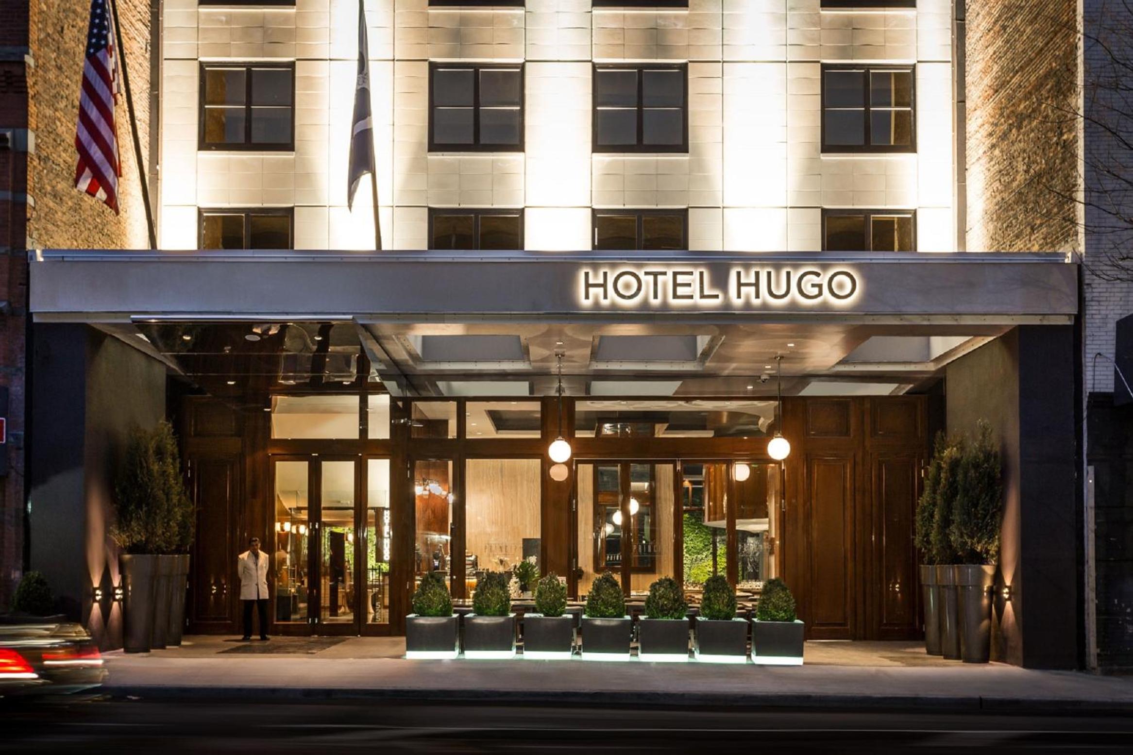 Illuminated street entrance and facade of Hotel Hugo at dusk, with modern glass doors, canopy, and potted shrubs.
