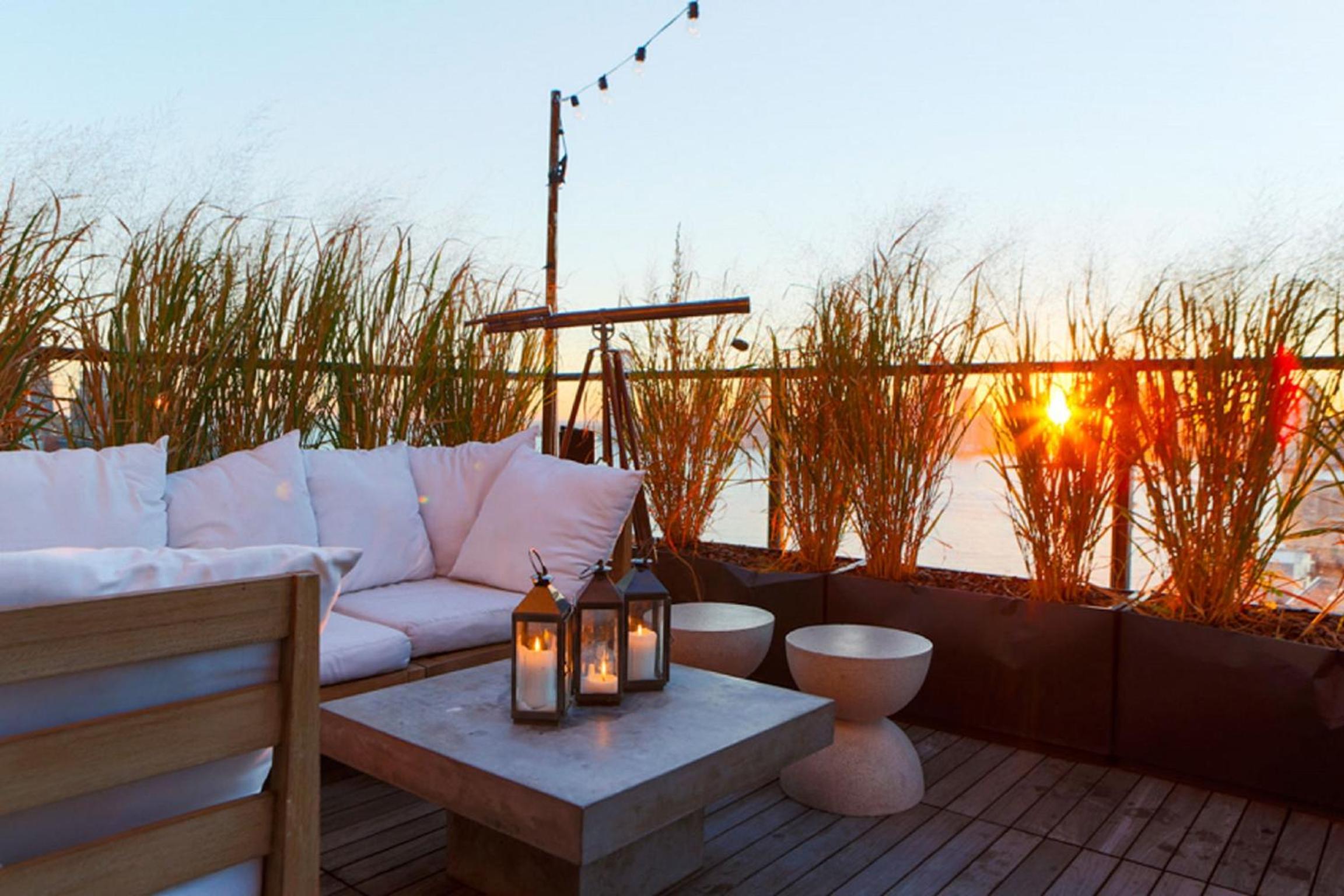Wooden rooftop deck with white cushioned seating, candle lanterns on a concrete table, tall grasses, and a sunset view.