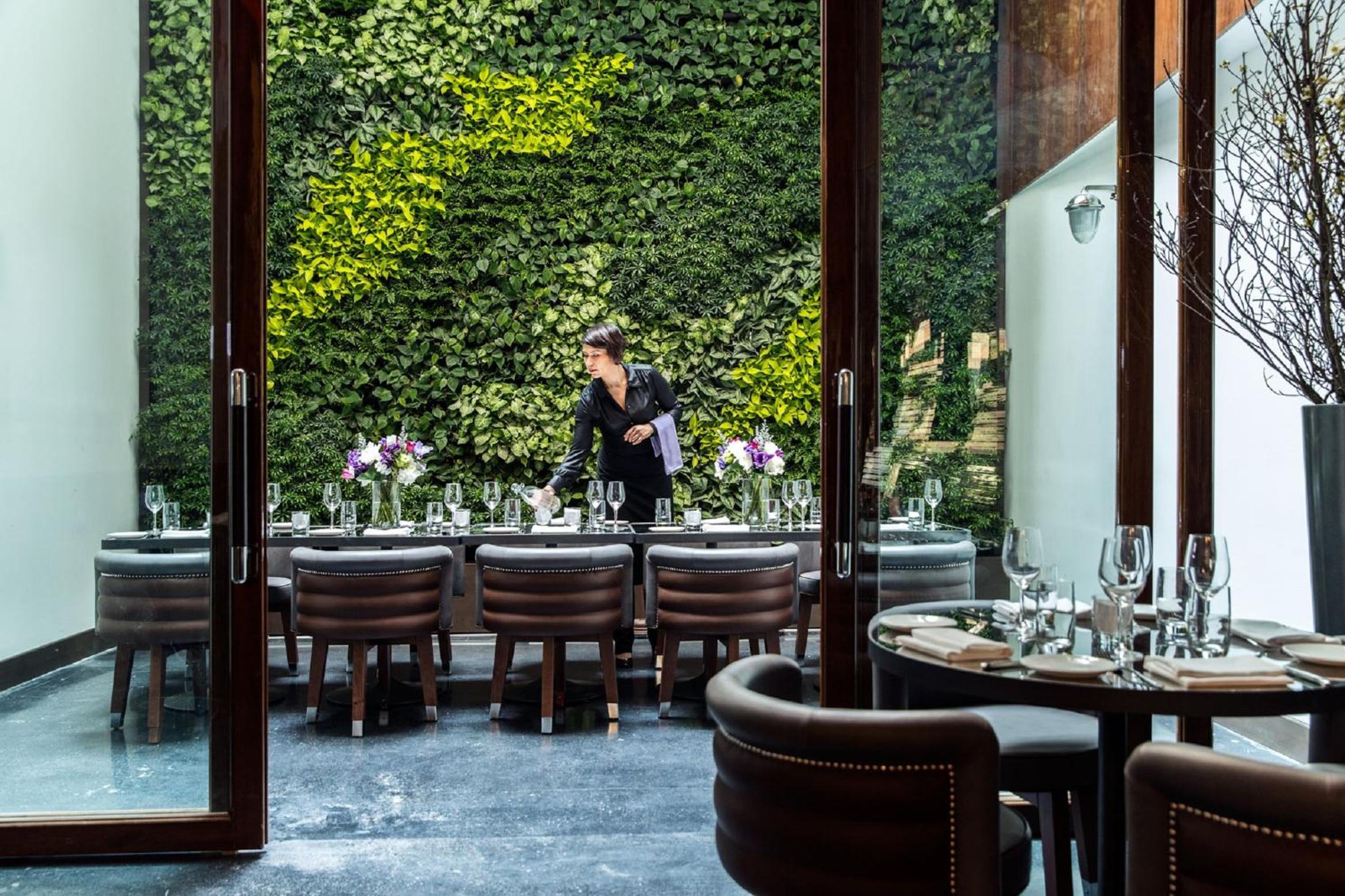 Restaurant dining space facing a vertical plant wall, with a server arranging stemware along set tables.