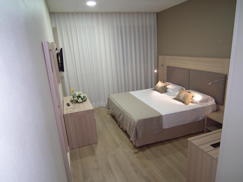 A bedroom with a neatly made bed, light wood furniture, sheer white curtains, and pale flooring.