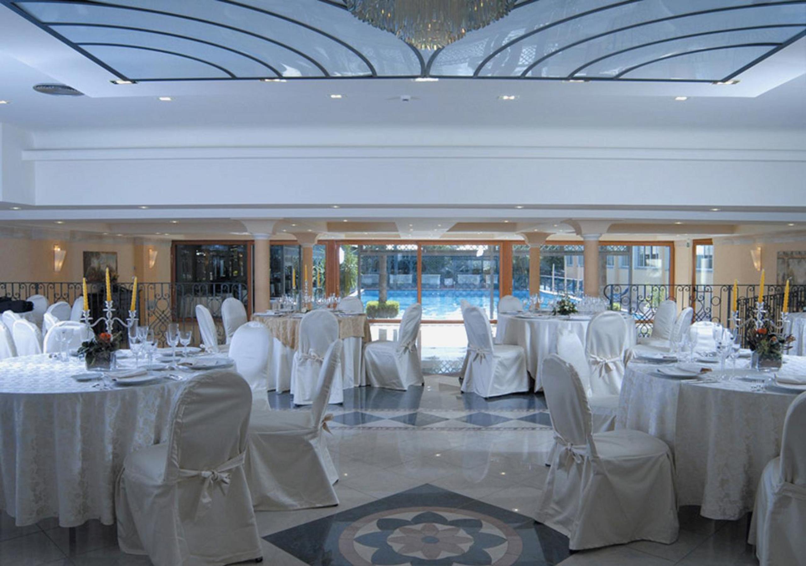 A banquet hall with round tables dressed in white linens and chair covers facing glass doors toward a pool.