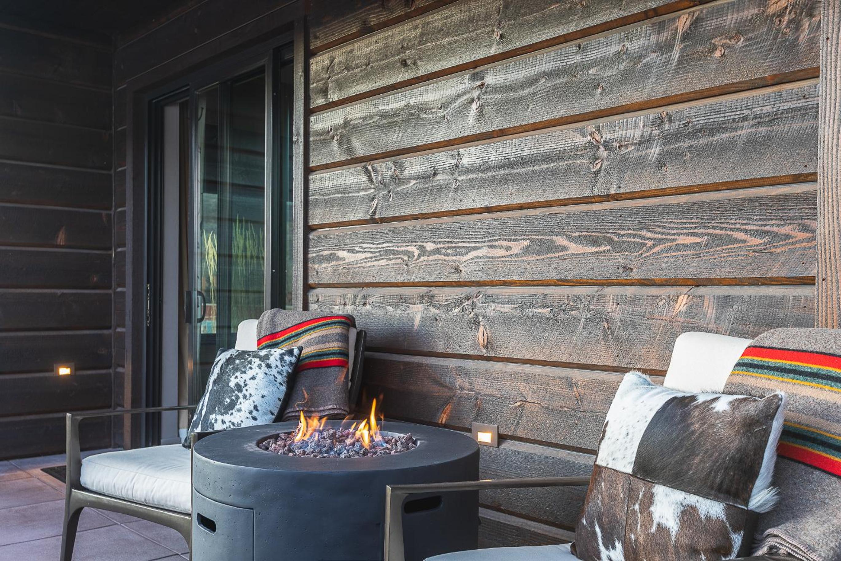 A balcony patio with weathered wood siding, armchairs, and a burning fire table.