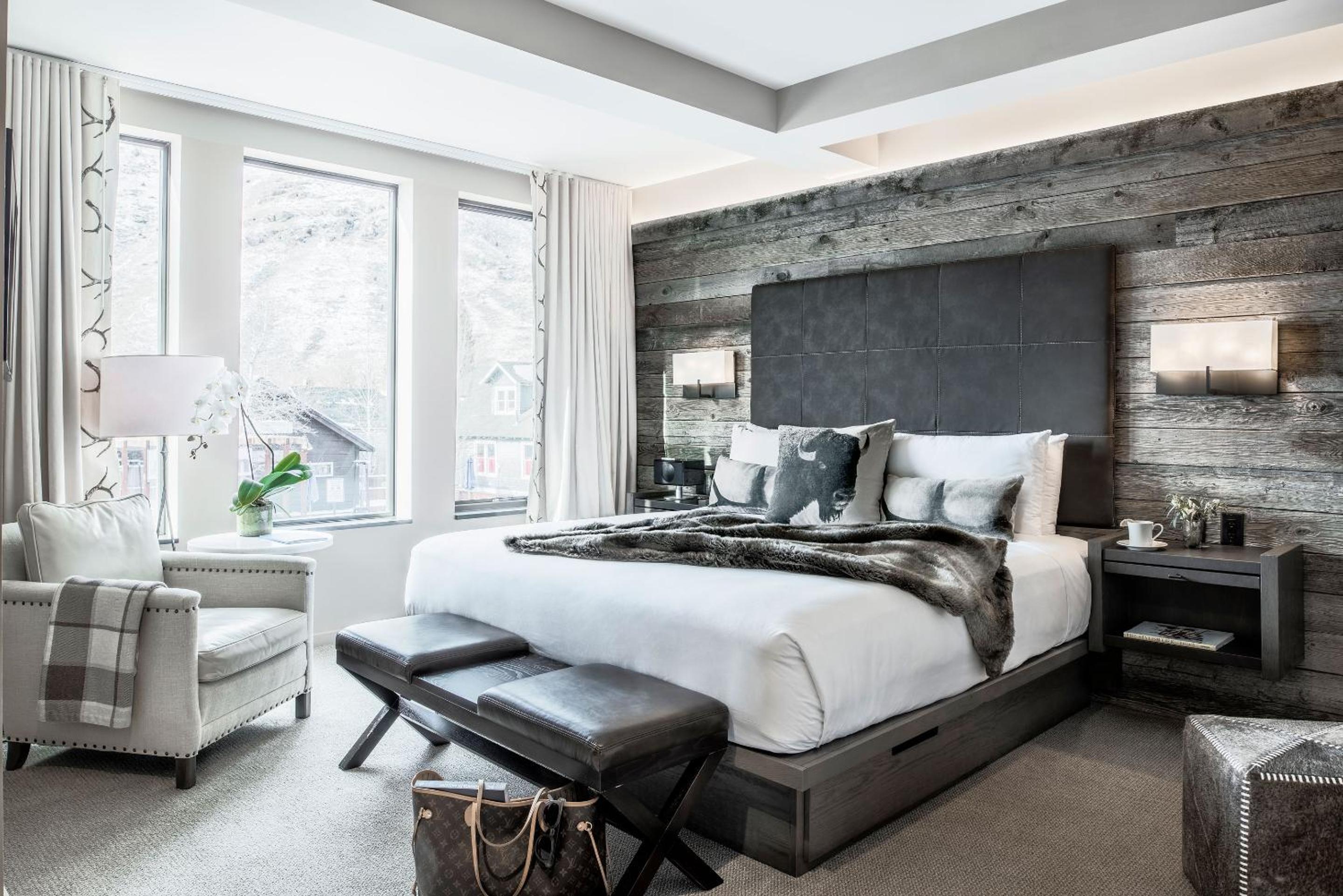 A bedroom with a leather headboard against reclaimed wood paneling, an armchair, and windows facing hills.