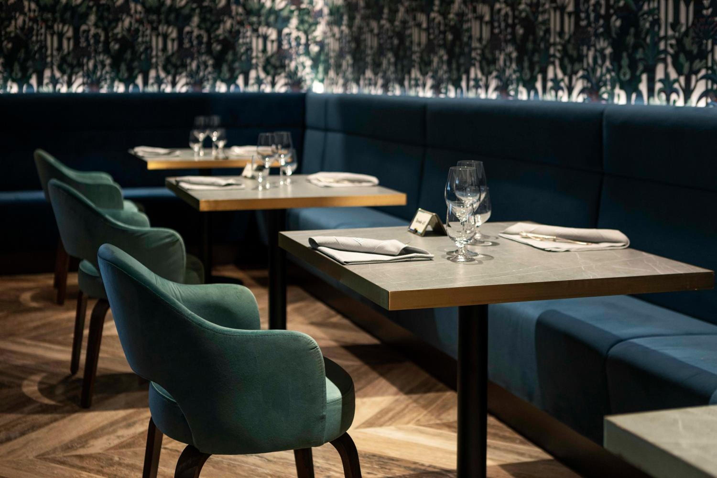 Restaurant dining area with plush teal armchairs, blue banquettes, and tables set with glasses and folded napkins.