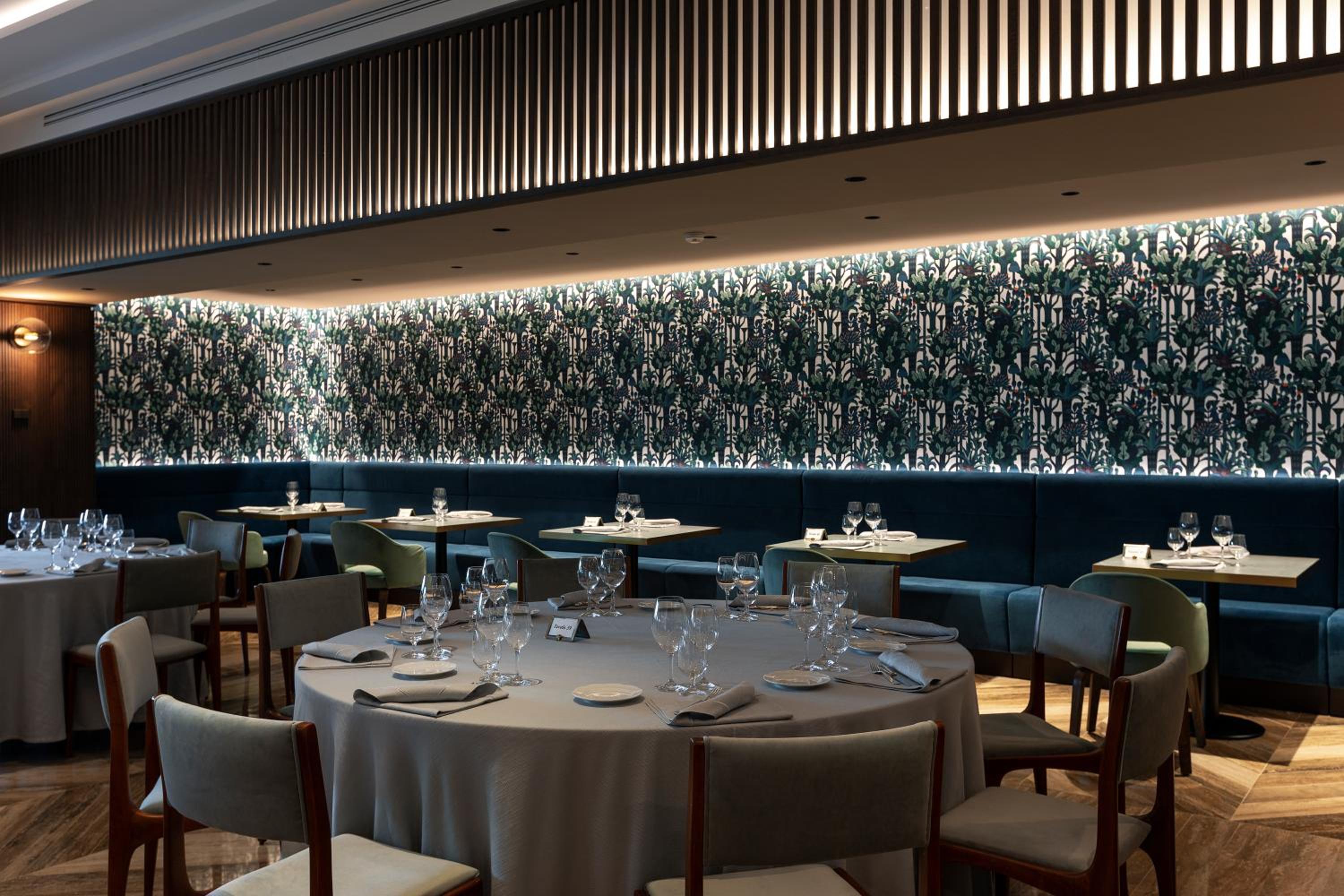 Spacious dining room with set round tables, wood-slatted ceiling fixtures, and patterned wallpaper under warm light.
