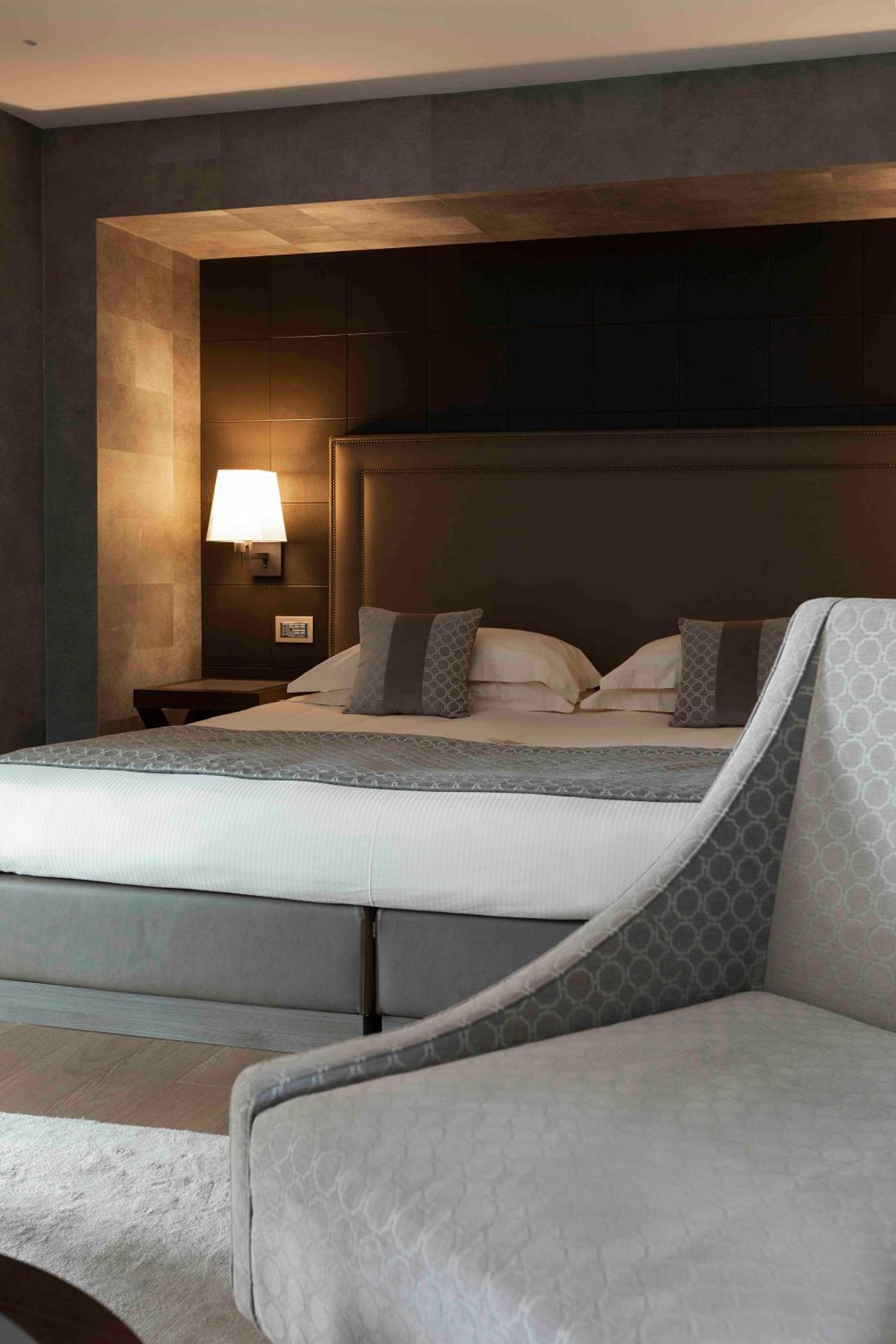 Hotel bedroom with an upholstered headboard, glowing bedside lamp, and a patterned armchair in the foreground.