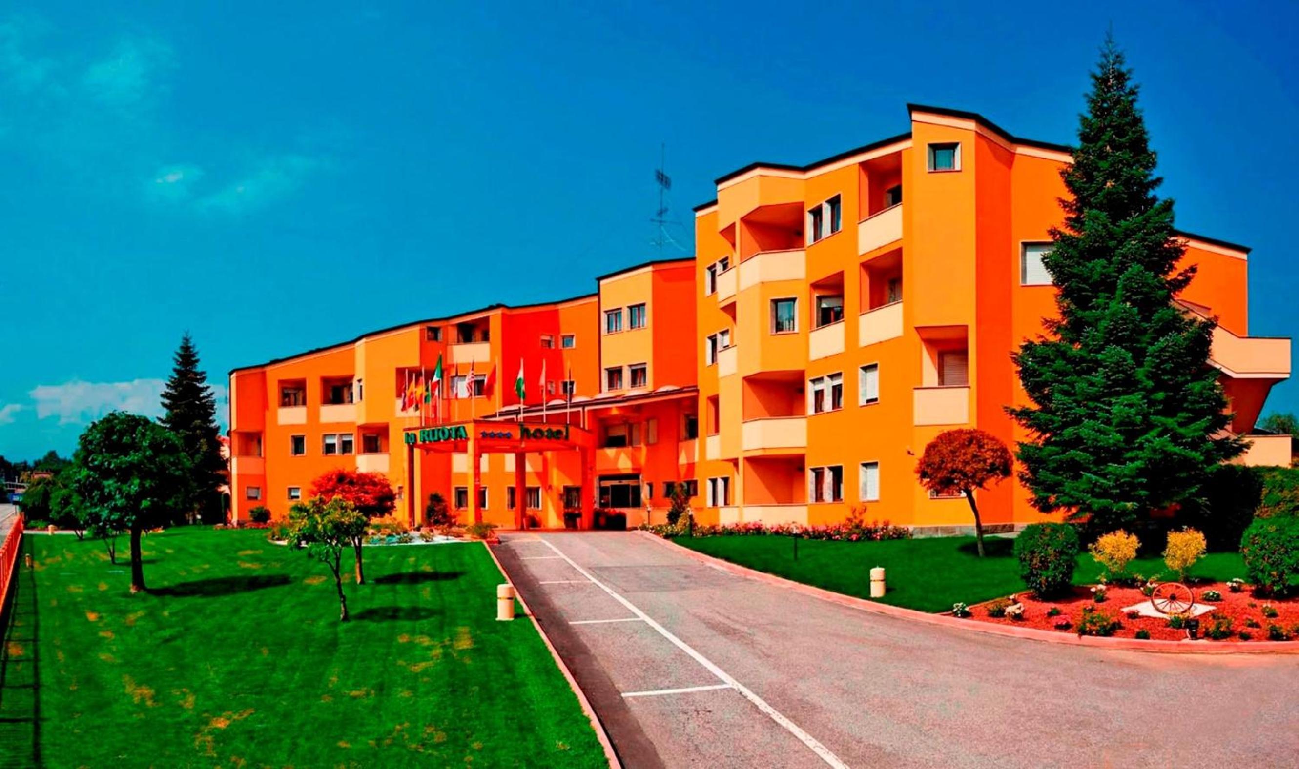Bright orange facade of Hotel La Ruota with balconies, a curved driveway, and green lawn under a blue sky.