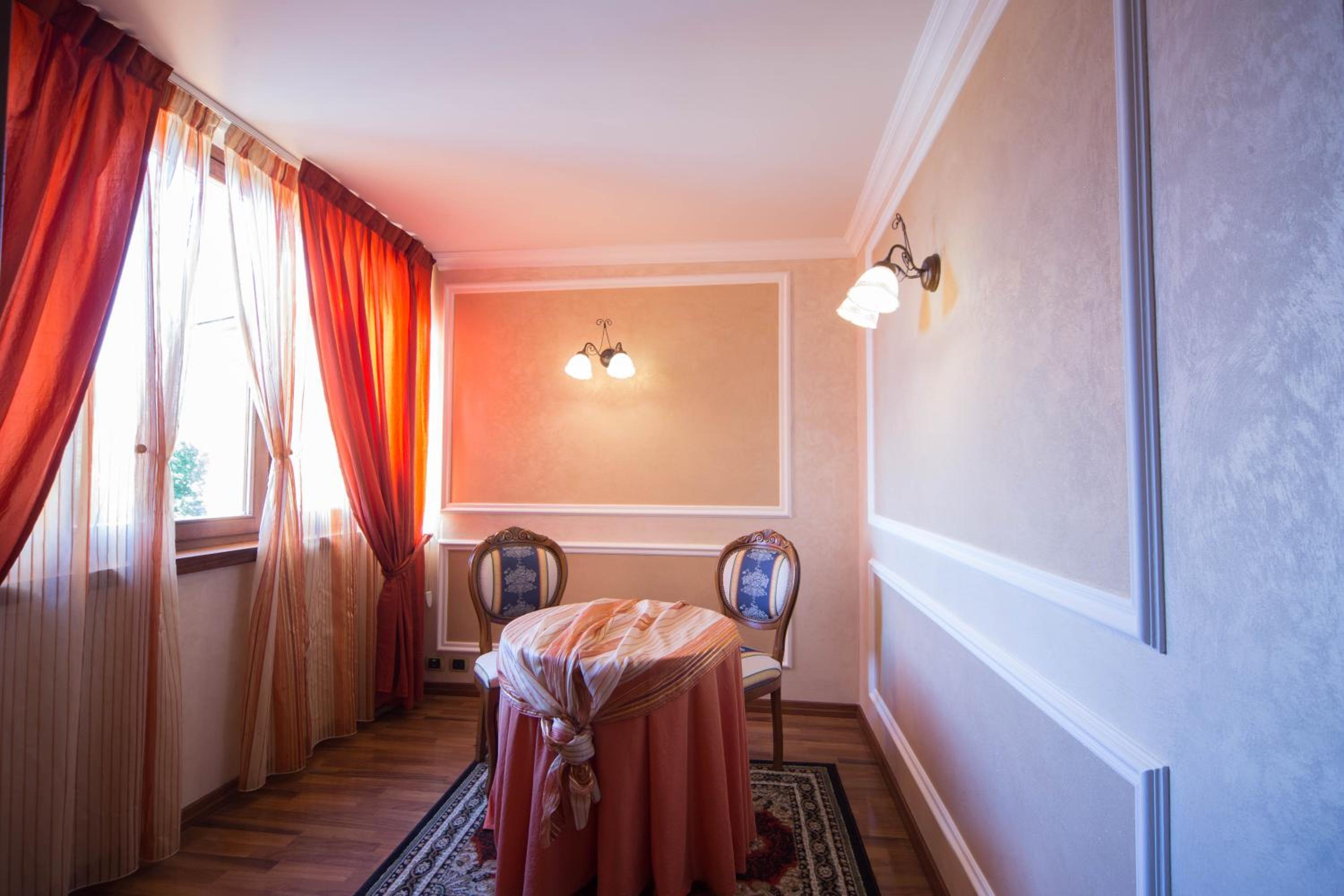 Hotel room alcove featuring draped orange curtains, a small covered table, ornate chairs, and wall sconces.