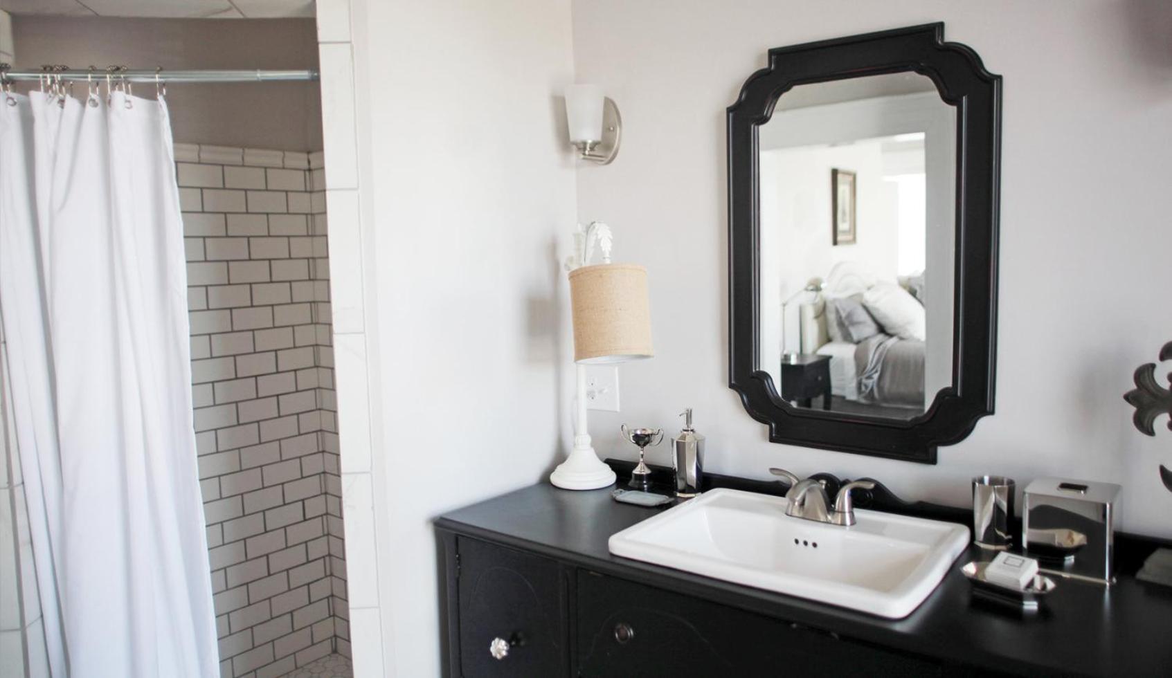 A black vanity with a white basin sits below a framed mirror next to a subway-tiled shower.
