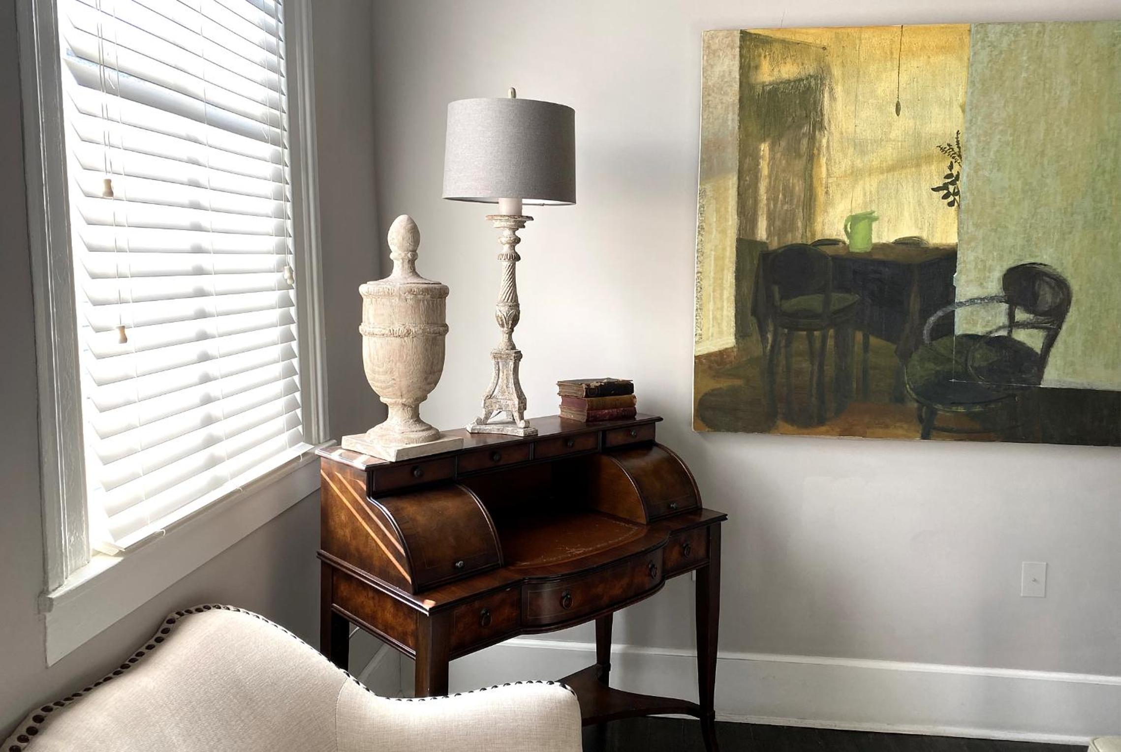 An antique rolltop desk with a table lamp and stone urn stands near a window beside a large painting.