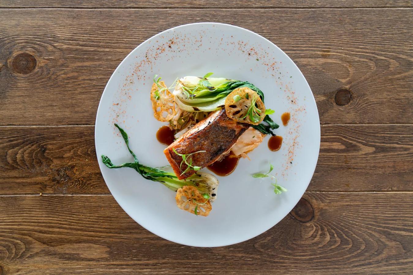 Plated seared fish with bok choy, crispy lotus root, and reduction sauce on a dark wood table seen from above.