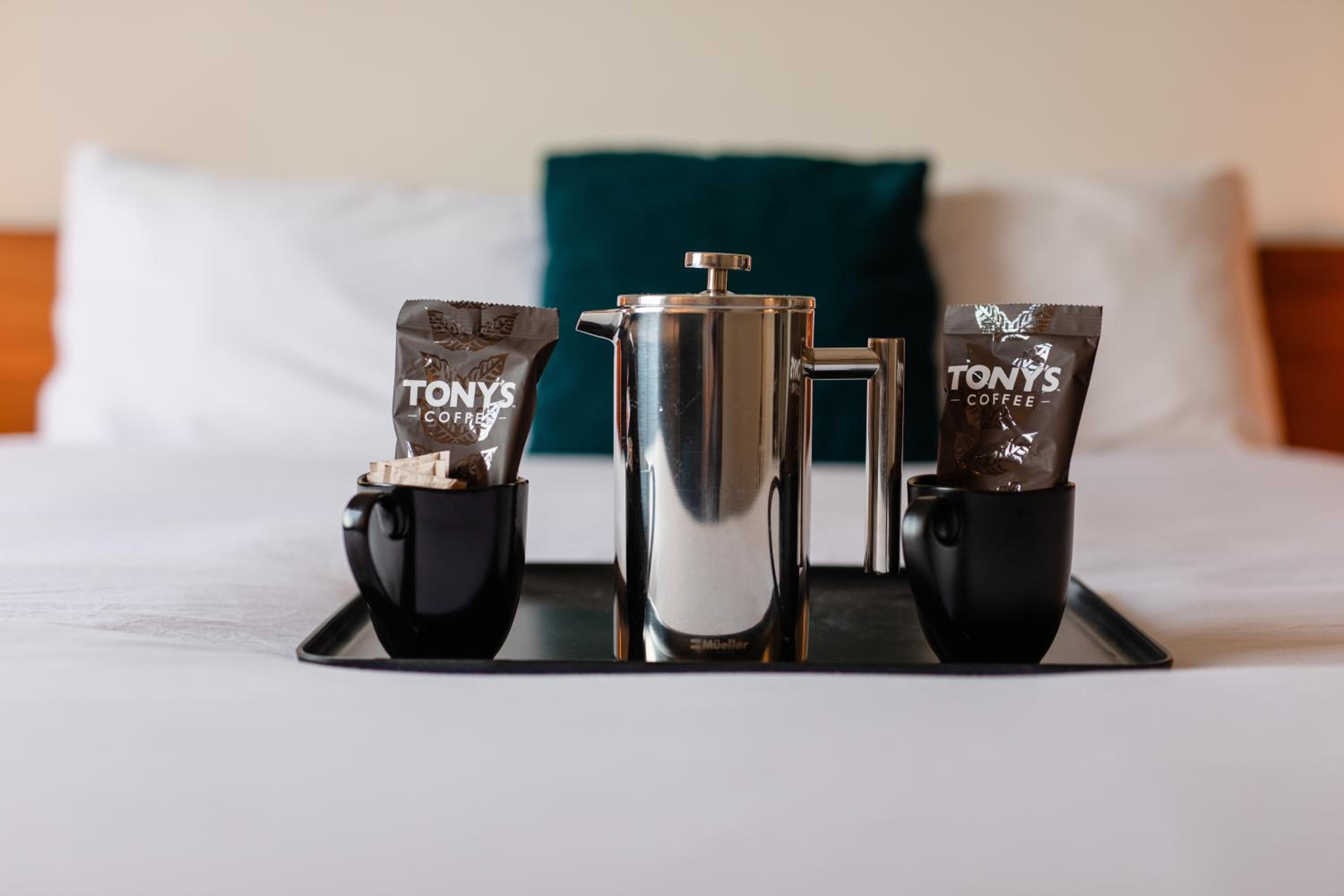 A metal French press and black mugs with coffee packets rest on a tray atop a bed.