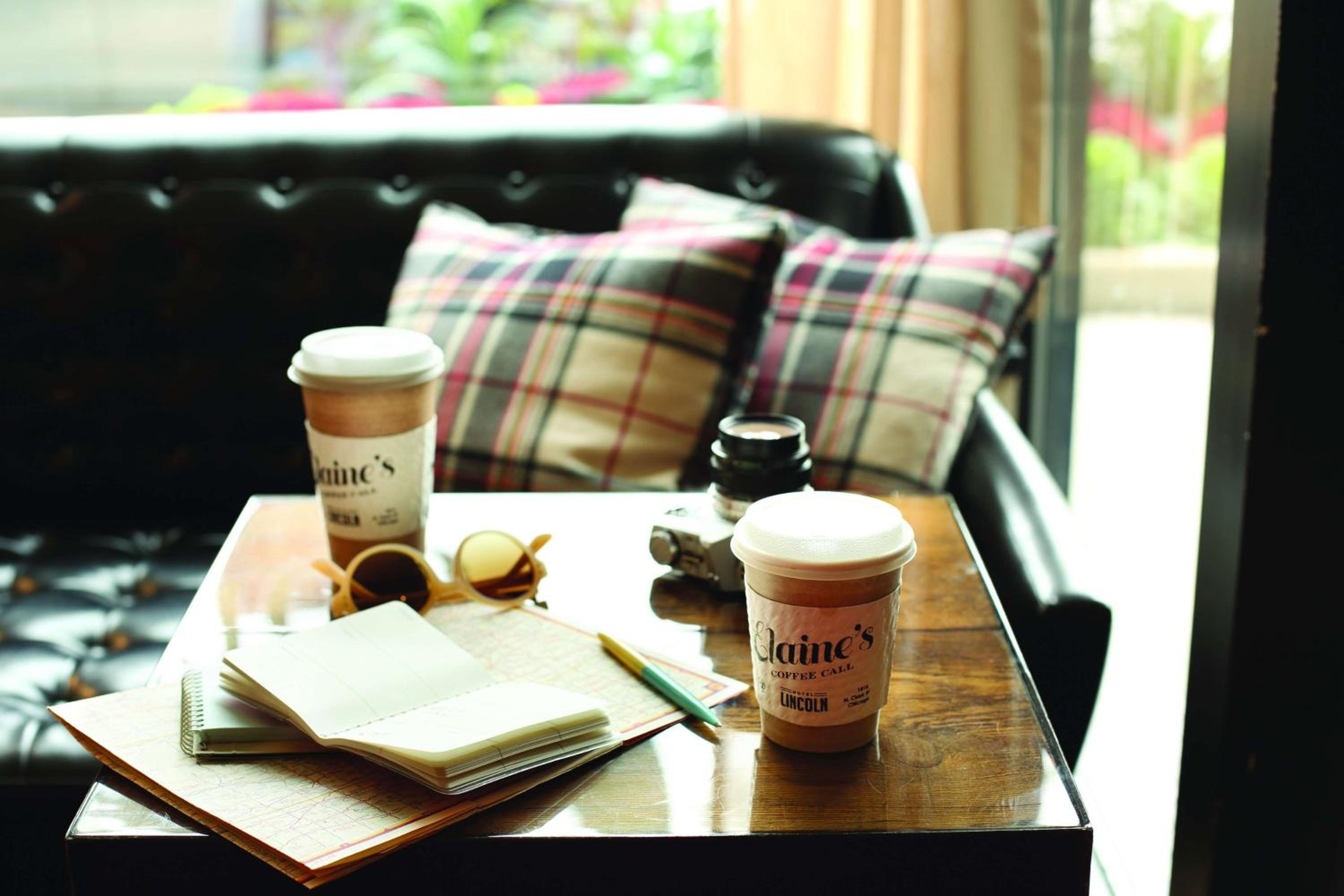 Coffee cups, a notebook, camera, and sunglasses on a low wooden table next to a leather sofa with plaid pillows.