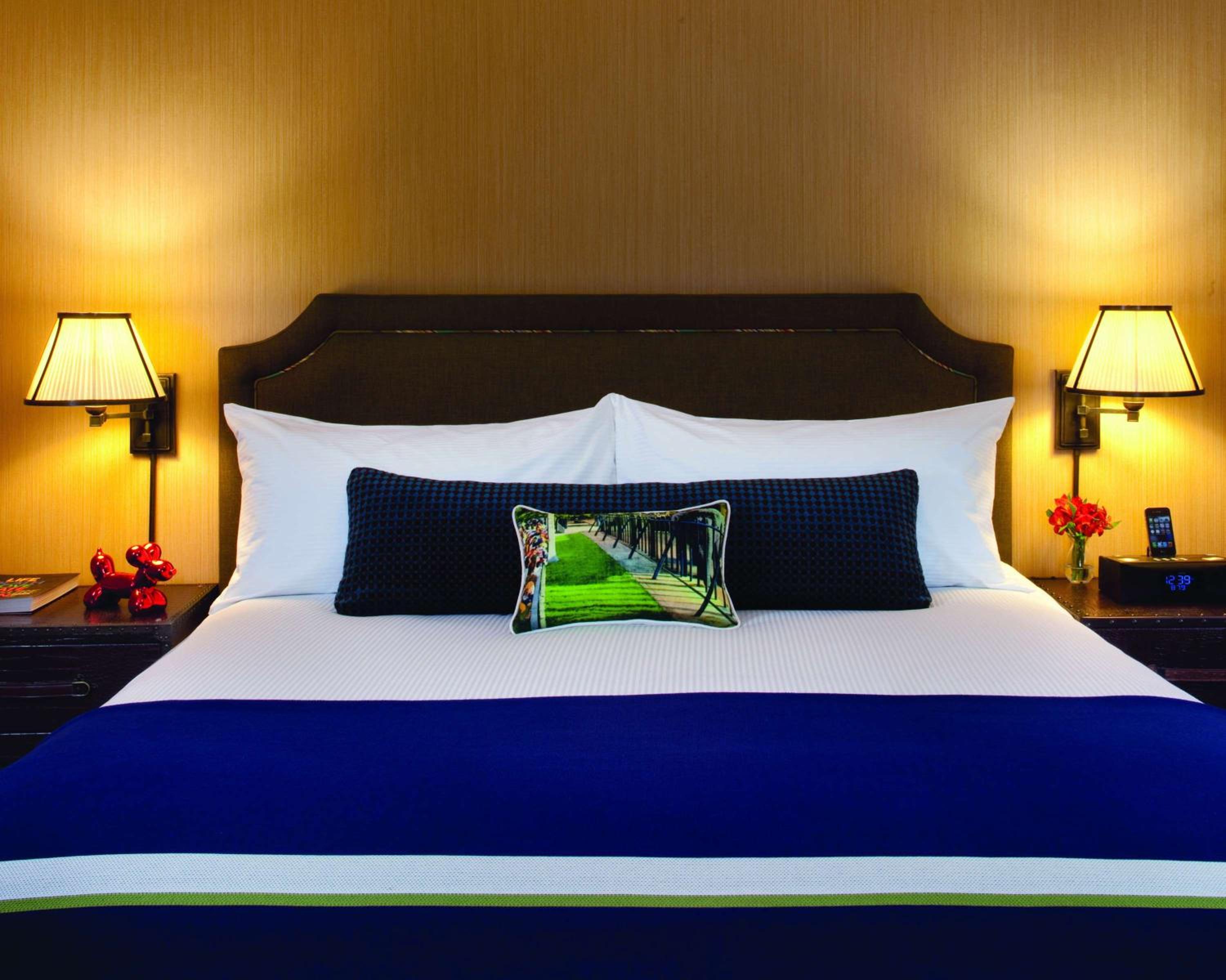 A hotel bed with crisp white linens, a blue throw blanket, and matching bedside lamps against textured wallcovering.