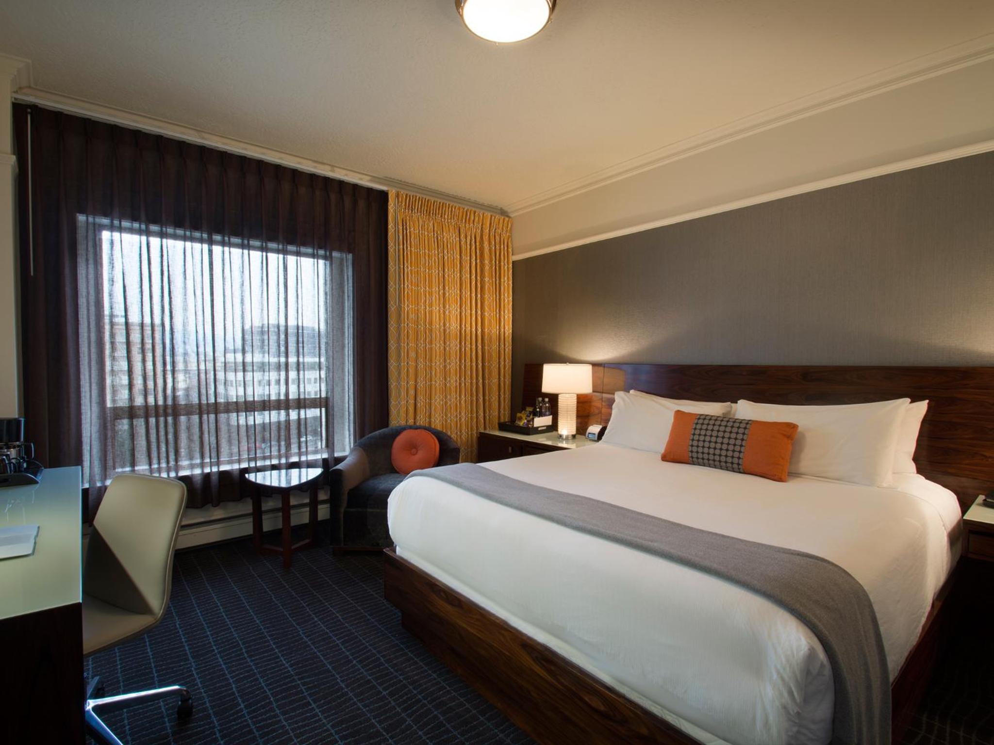 A hotel bedroom with a wood-framed bed, white linens, patterned carpet, and sheer curtains by the window.
