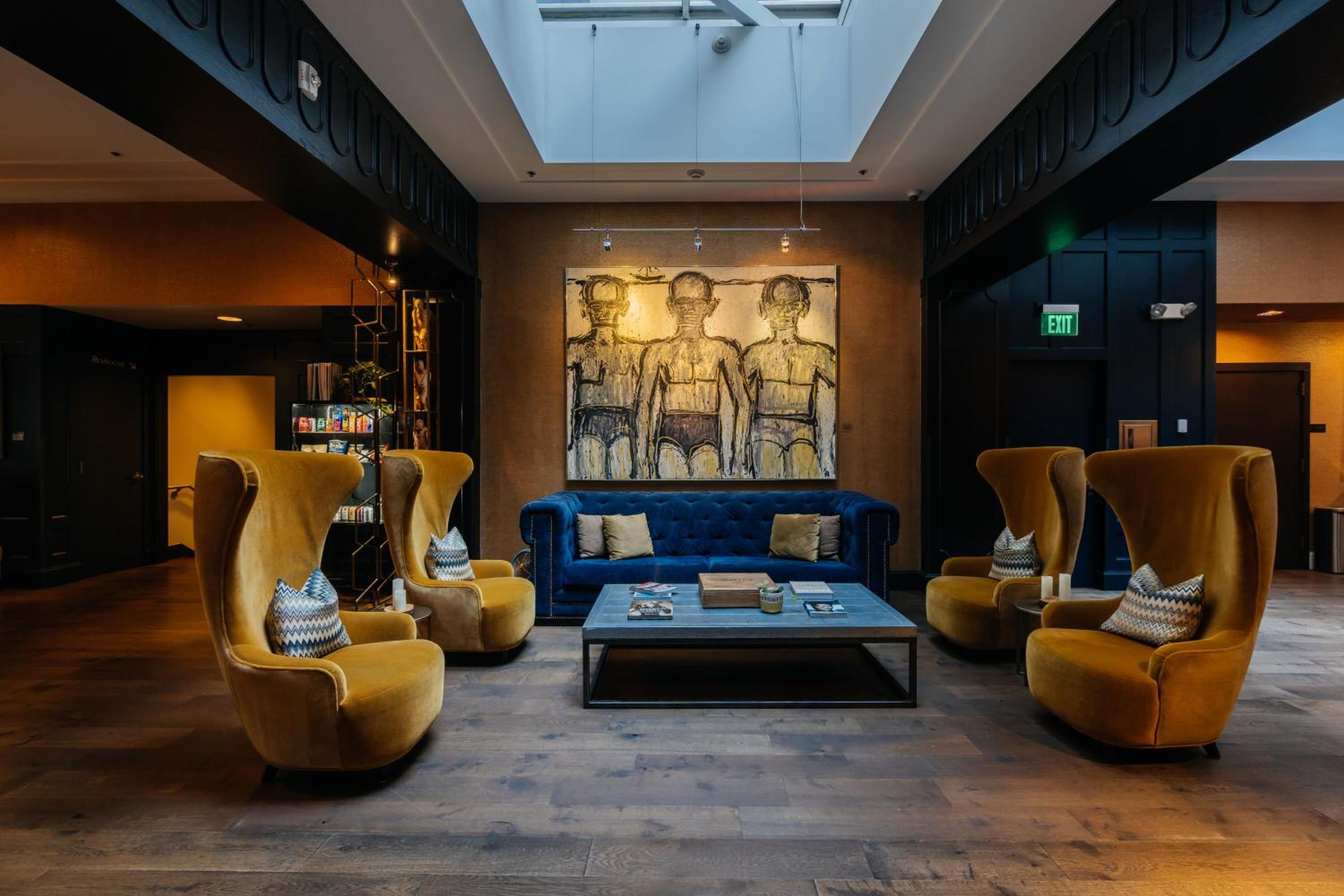 A lounge area beneath a skylight featuring tall yellow armchairs, a blue tufted sofa, and modern wall art.