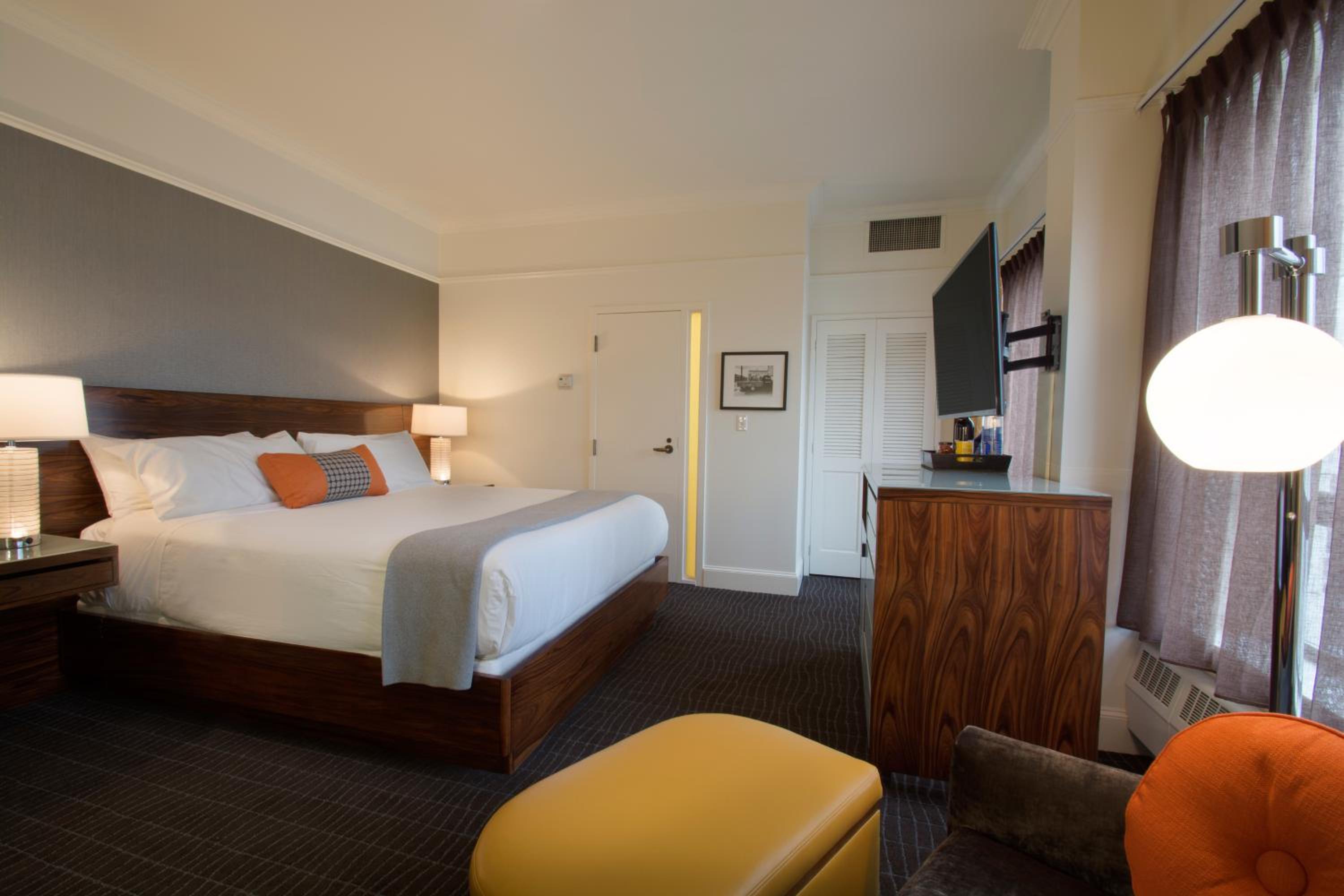 A guest room with a wooden bed frame, gray accent wall, mounted television, and a mustard yellow footstool.